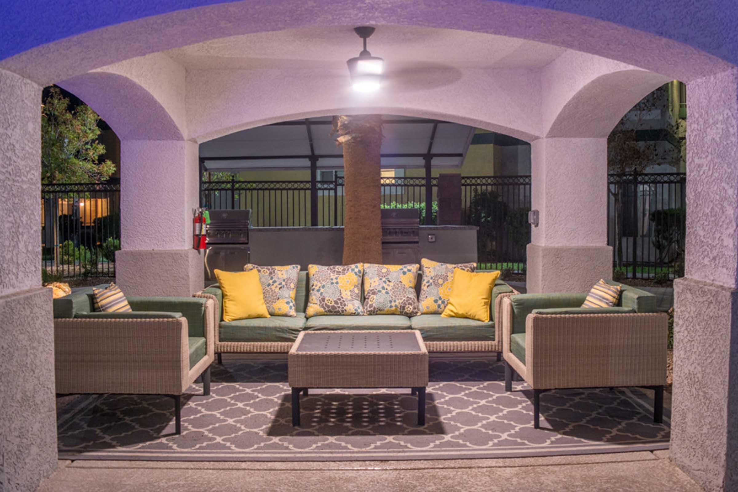 Cozy outdoor seating area under an archway, featuring a green sofa with yellow and floral accent pillows, surrounded by two matching chairs. A coffee table sits in the center on a patterned rug, illuminated by a hanging light. The setting is peaceful, suggesting a relaxing ambiance at night.