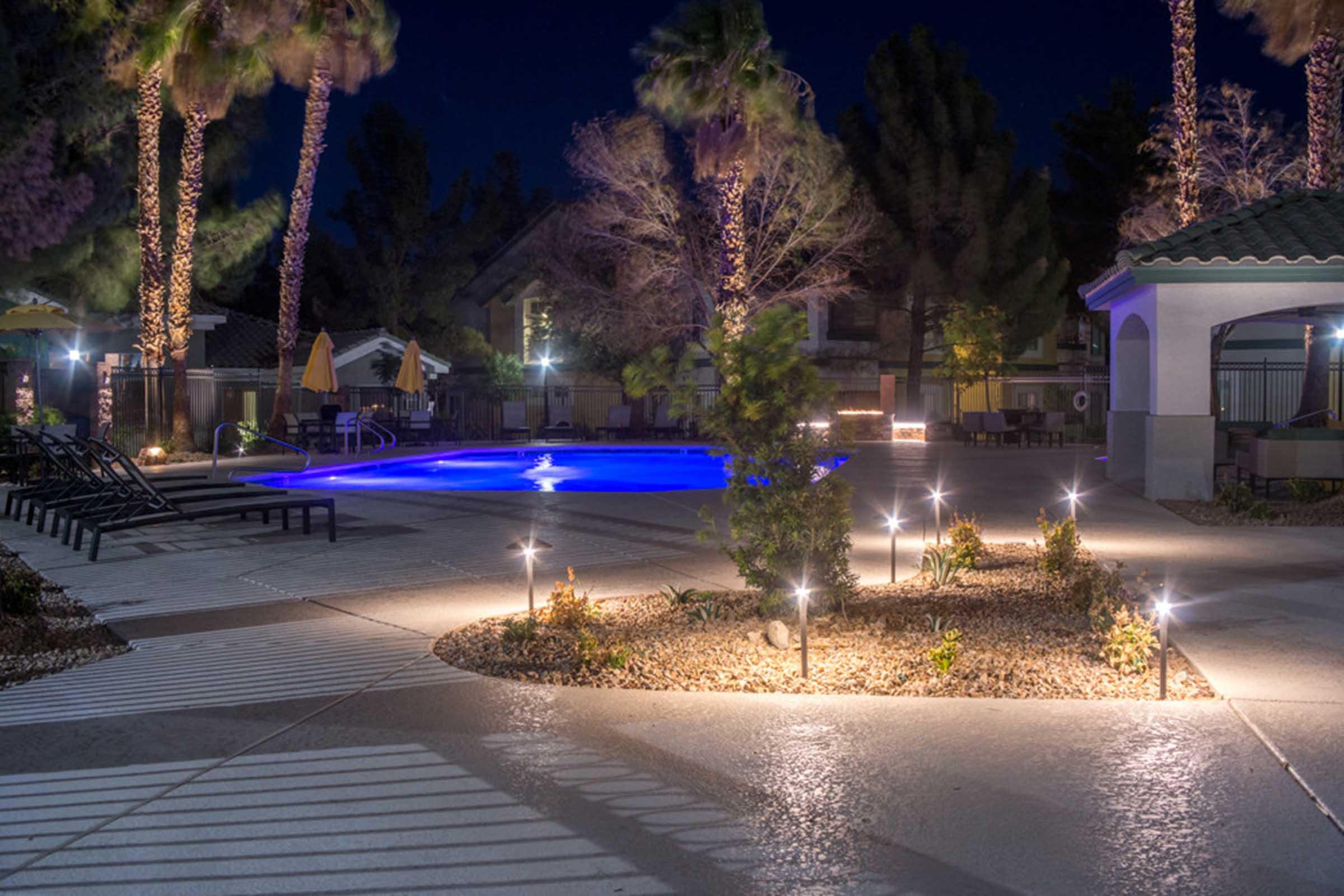 A nighttime view of a swimming pool area surrounded by palm trees. The pool is illuminated with a soft blue light, and the surrounding landscape features well-lit pathways, decorative plants, and lounge chairs. The ambiance is tranquil and inviting, with buildings visible in the background.