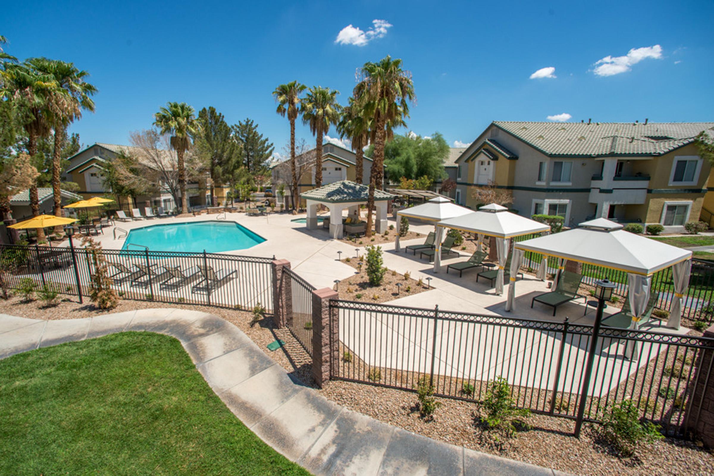 A spacious outdoor pool area surrounded by palm trees and lounge chairs, featuring white cabanas. In the background, there are residential buildings with a clear blue sky overhead. The area is well-maintained with landscaped gardens and a gated fence, creating a relaxing atmosphere for leisure activities.