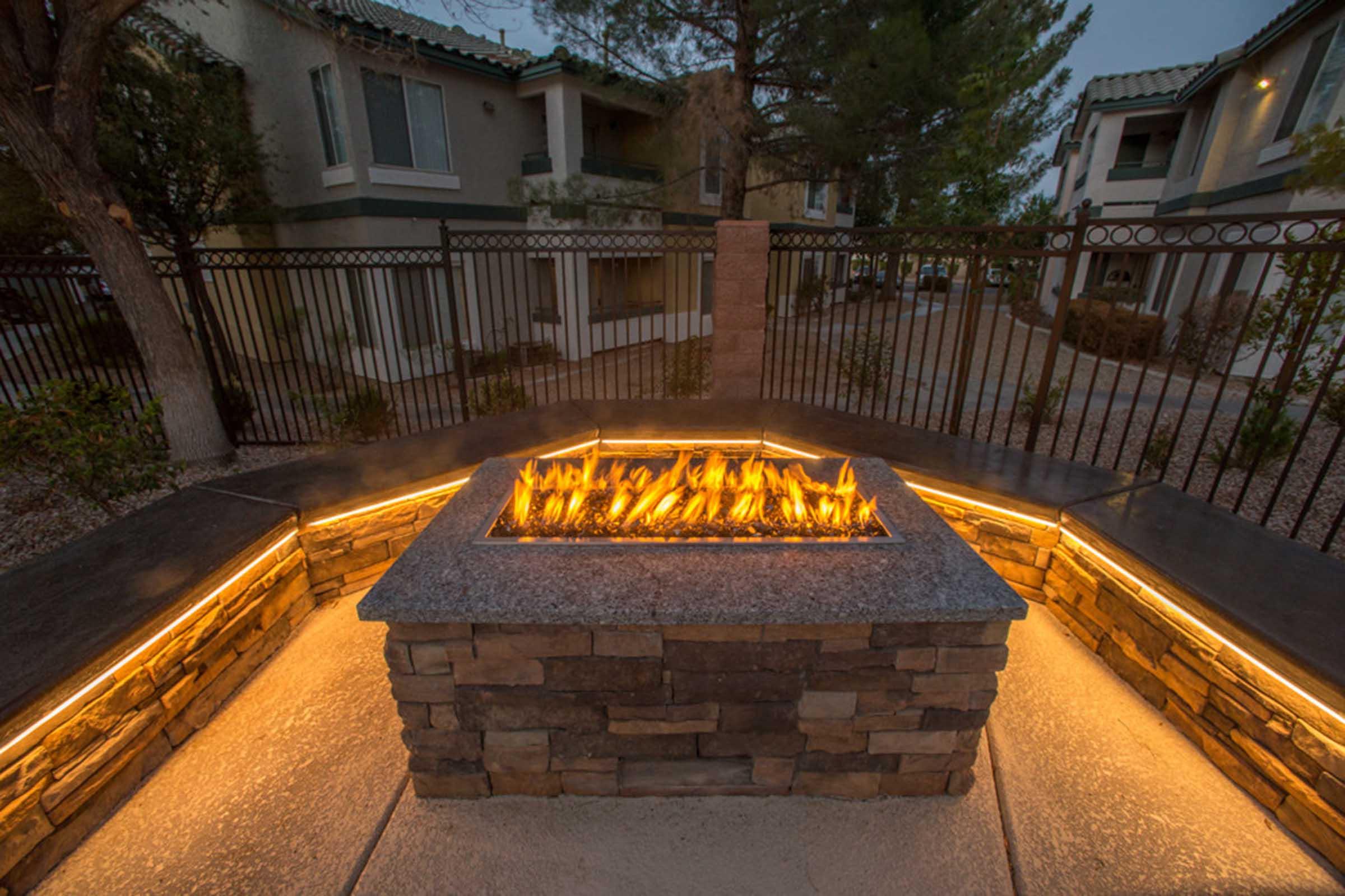 A cozy outdoor fire pit surrounded by stone and concrete seating, illuminated by soft lighting. Nearby are green shrubs and a backdrop of residential buildings, creating a warm and inviting atmosphere in the evening.