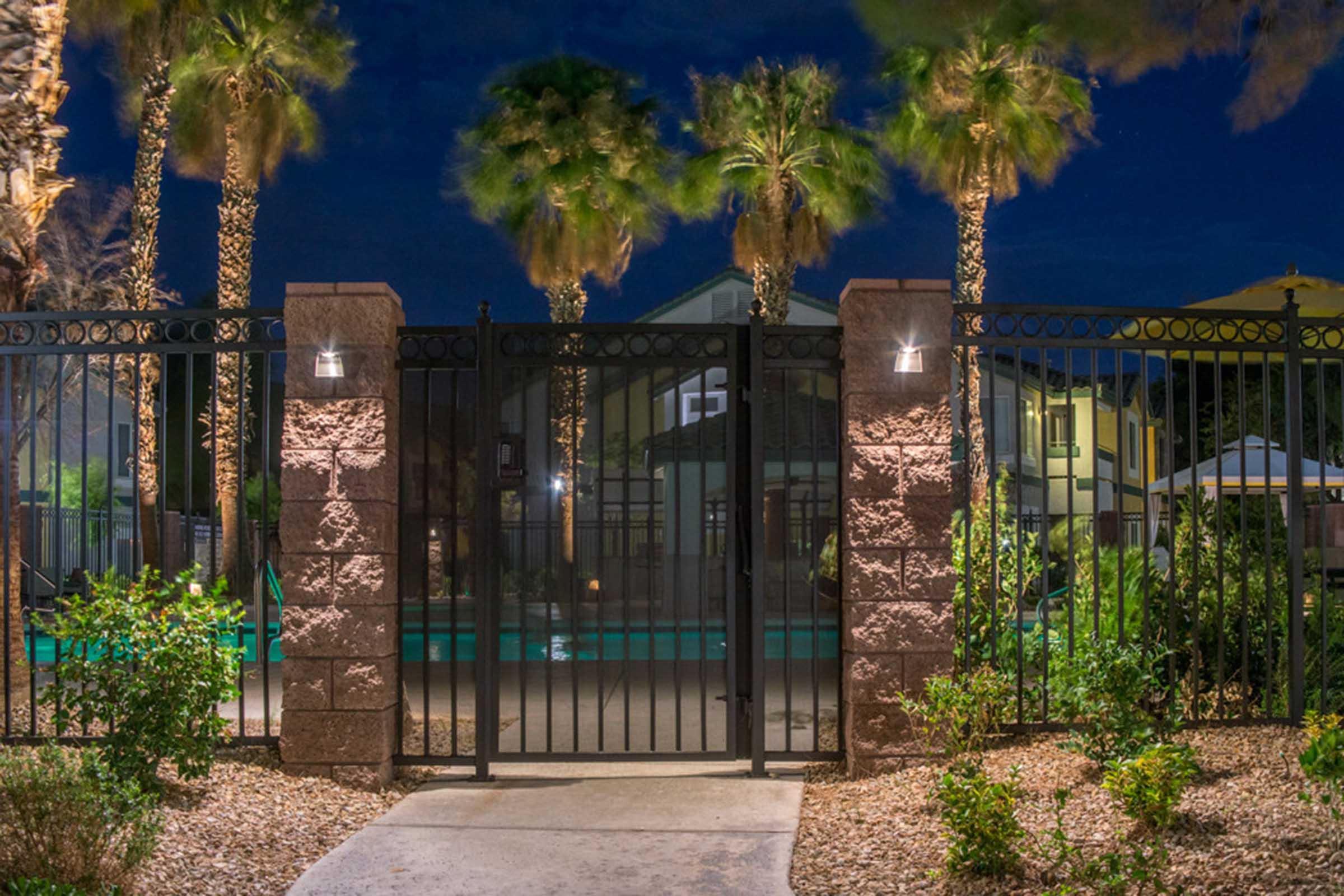 A gated entrance to a pool area at night, surrounded by palm trees and landscaped shrubs. The lighting highlights the gate, and a glimpse of the pool is visible in the background, creating a serene atmosphere.