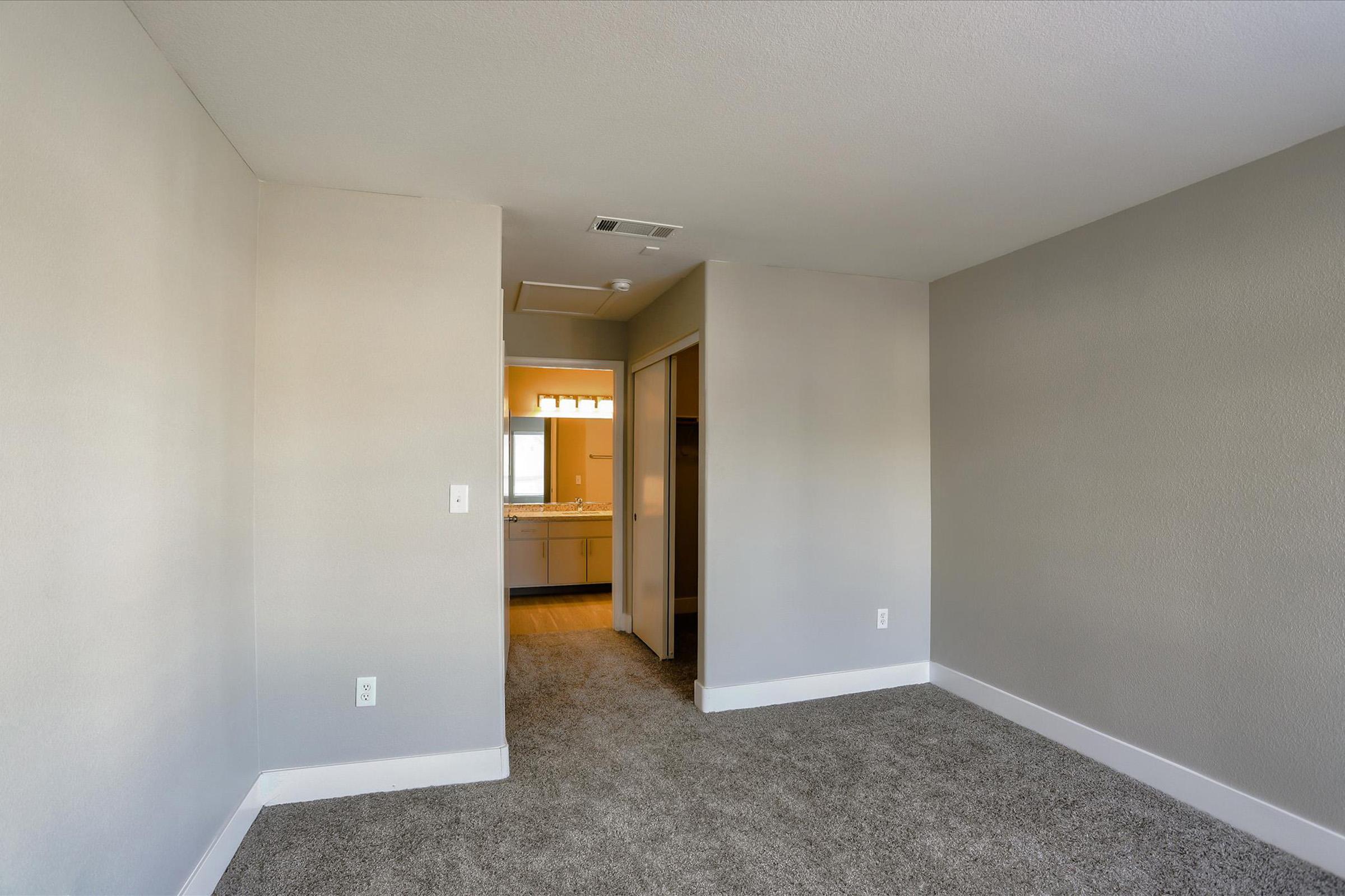 A spacious and empty room with light gray walls and carpeted flooring. On the left, there is a doorway leading to a bathroom area, which has a sink visible in the background. Natural light enters through a window, creating a bright atmosphere in the space. The room is ready for furnishing.