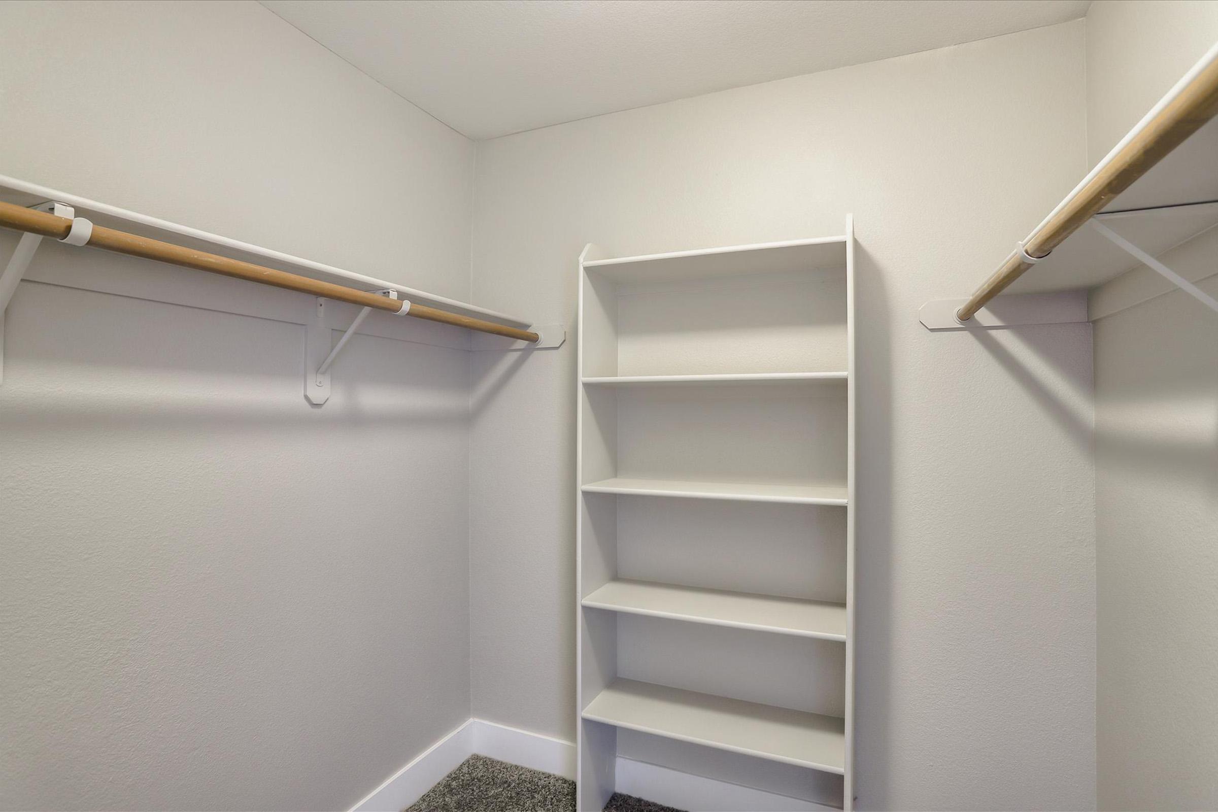 Empty closet space with light gray walls, featuring two coat rods on the sides and a white shelf unit in the center. The floor is dark and textured, providing a clean and organized appearance suitable for storage.