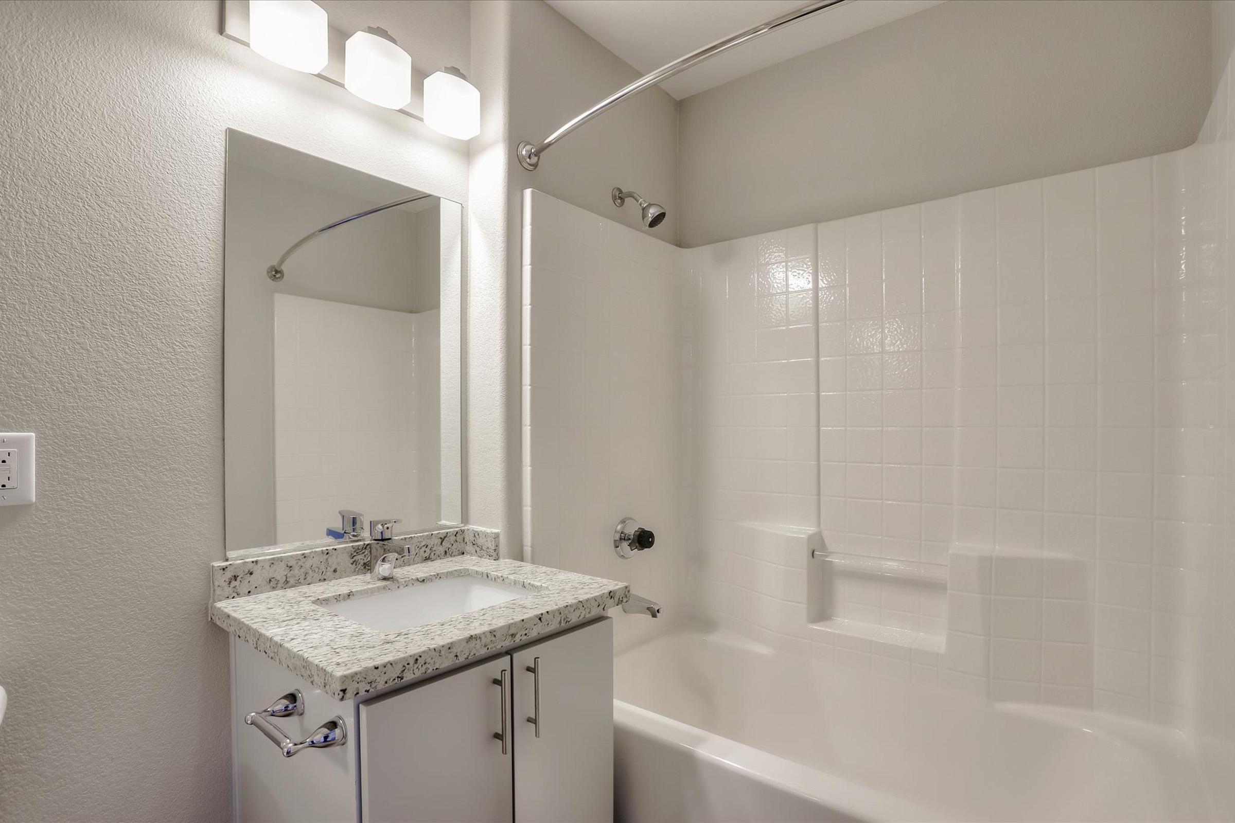A clean bathroom with a white tiled shower and bathtub. There is a modern sink with a granite countertop and a mirror above it. The walls are painted in a light color, and there are four light fixtures above the mirror. The overall appearance is bright and contemporary.