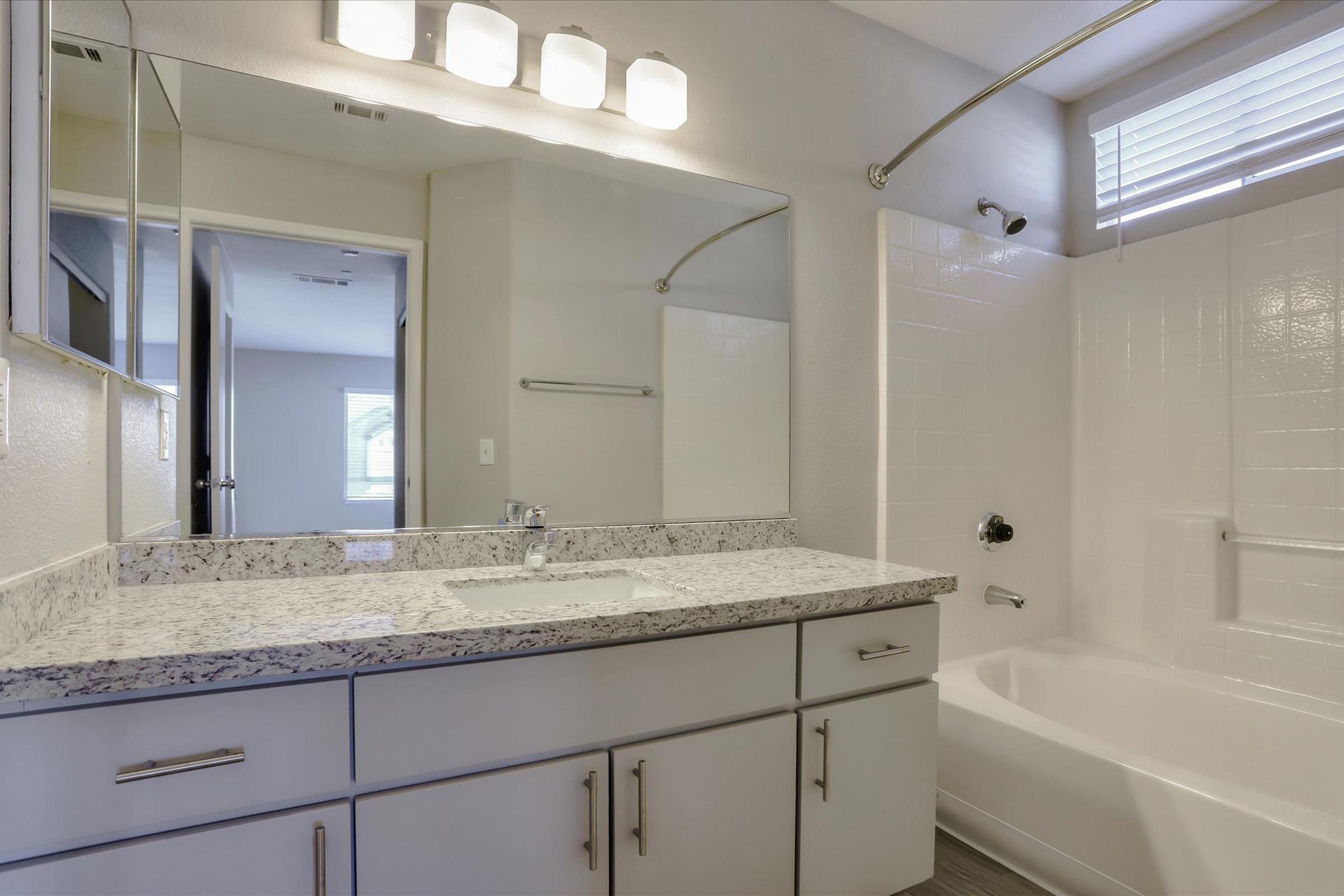 A modern bathroom featuring a white bathtub, a granite countertop with a sink, and a mirror above the sink. The walls are painted light colors, and there is a window with blinds for natural light. The lighting fixtures above the mirror provide additional illumination. The floor is finished with a light-colored material.