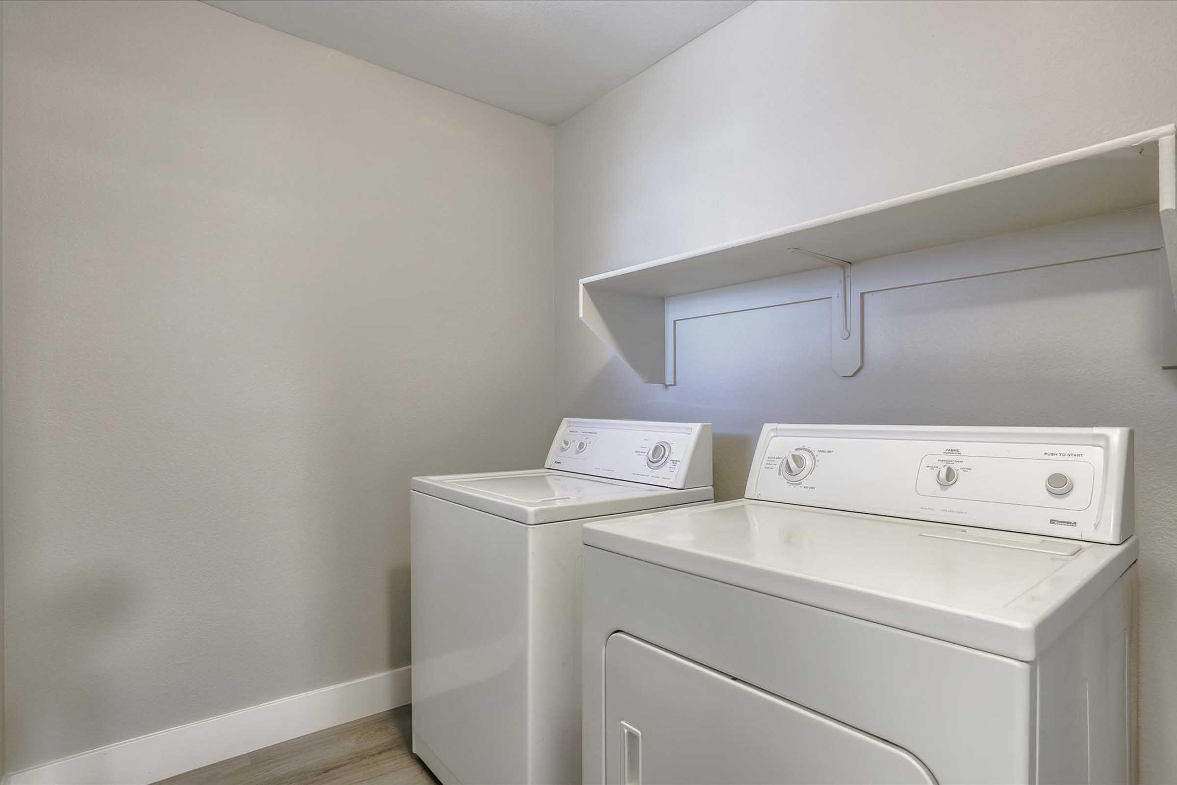 A clean and modern laundry room featuring two white appliances: a washing machine on the left and a dryer on the right. The walls are light gray, and a shelf above provides storage space. The flooring is a light-colored laminate, creating a bright and inviting atmosphere.