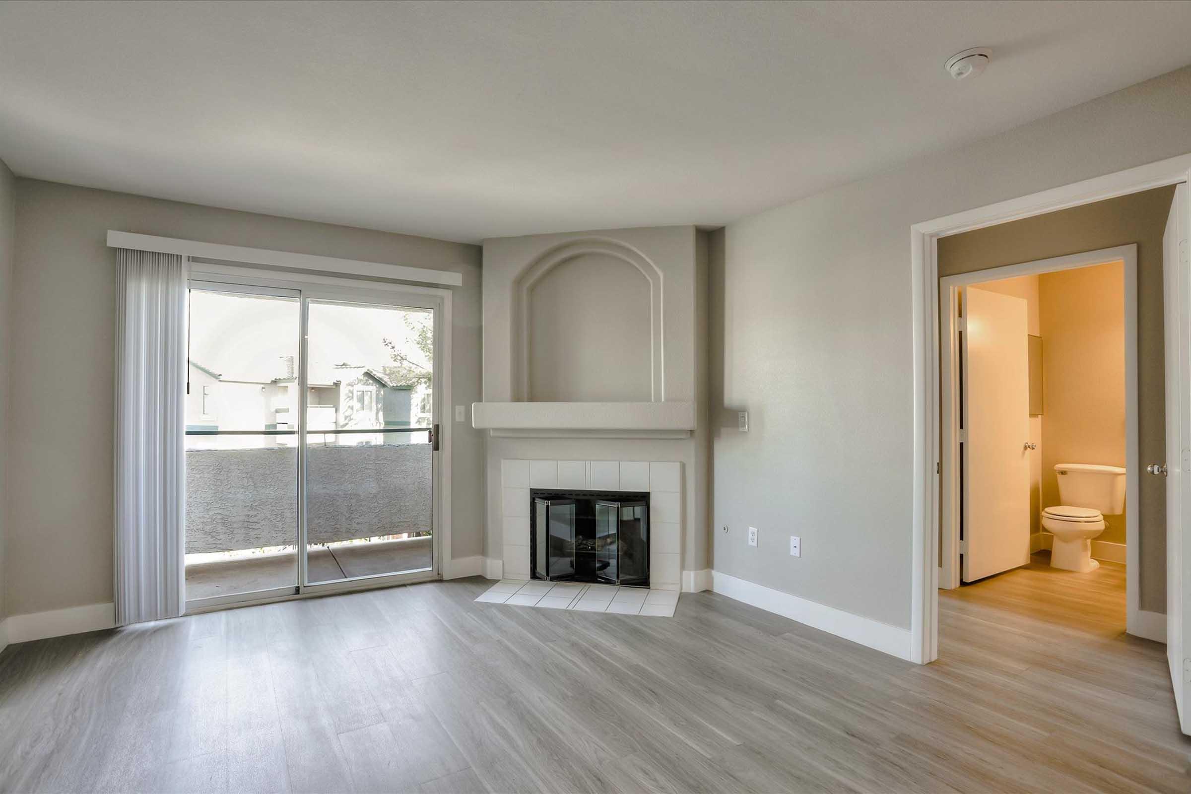 A spacious living room featuring a fireplace, large sliding glass doors leading to a balcony, and light-colored walls. A doorway on the right leads to a bathroom with a toilet. The space has modern flooring and ample natural light.