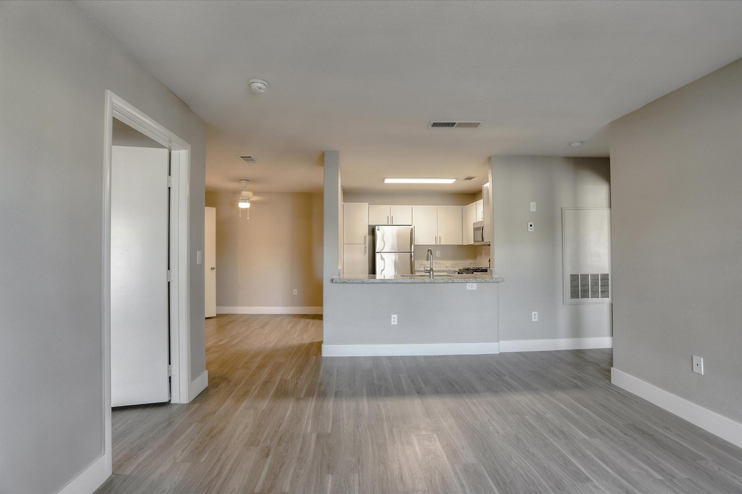 Interior view of a modern, open-concept kitchen and living area. The space features light gray walls, wood-look flooring, a kitchen island with a countertop, stainless steel appliances, and a ceiling fan. A doorway leads to another room, enhancing the spacious feel of the layout.