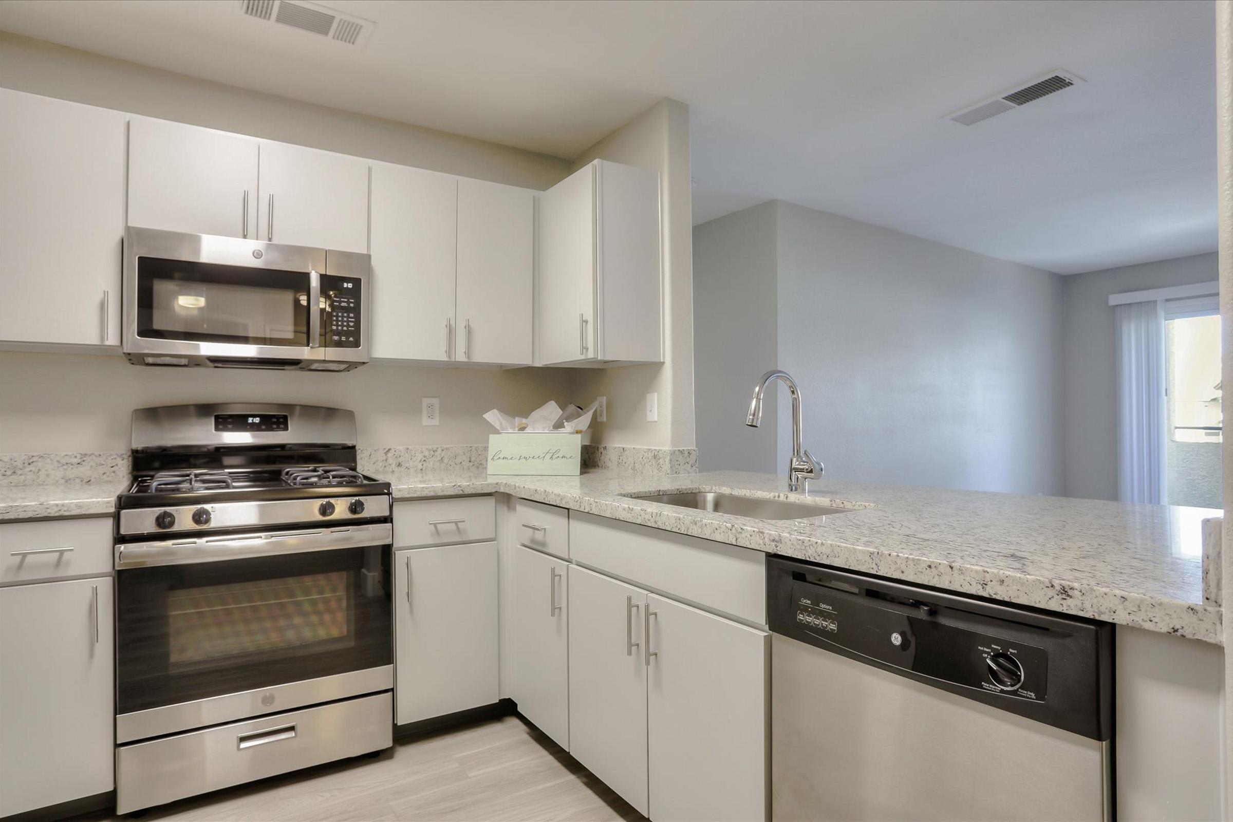 Modern kitchen featuring stainless steel appliances, including a microwave and a gas stove. The cabinets are sleek and white, complemented by a granite countertop. There is a sink with a modern faucet and a dishwasher. Natural light illuminates the space, highlighting the clean and contemporary design.