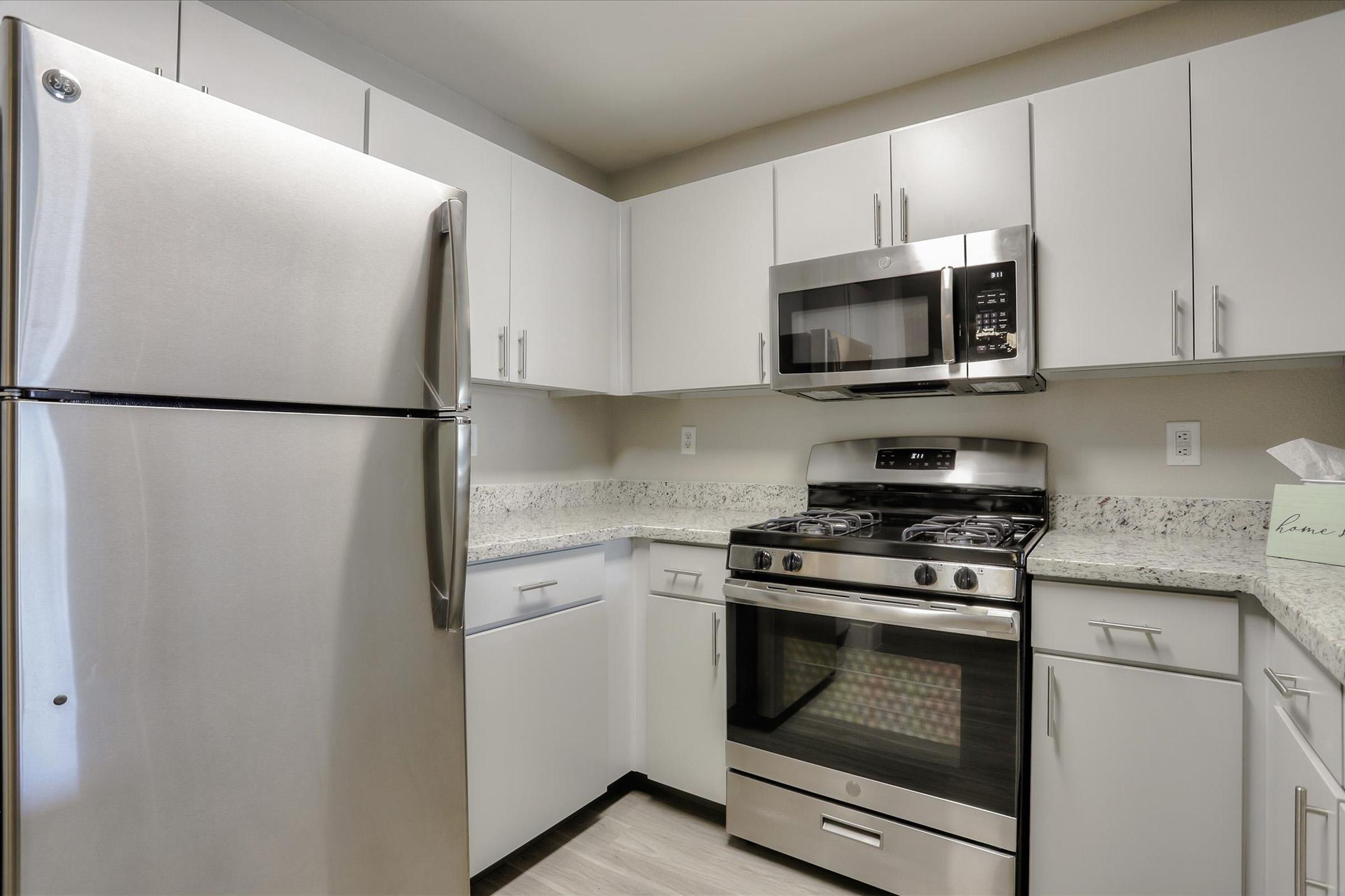 A modern kitchen featuring stainless steel appliances, including a refrigerator, microwave, and a gas stove with an oven. The cabinets are white, and the countertops are a light-colored granite, providing a clean and contemporary look. The floor is light-colored, complementing the overall design.