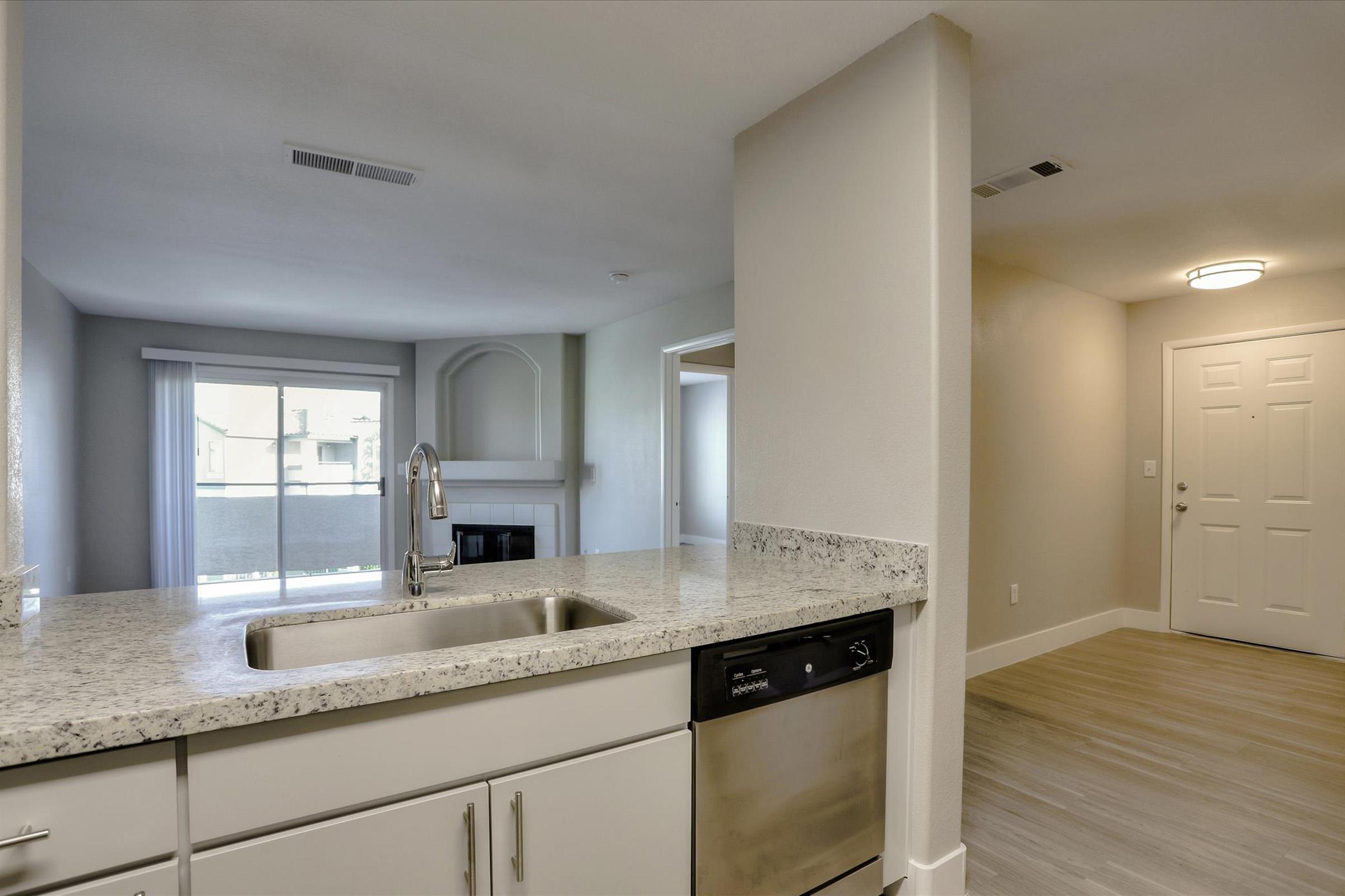 A modern kitchen with a granite countertop and stainless steel dishwasher, overlooking a spacious living area with large windows. The living room features a fireplace and entryway to a hallway with a closed door. The overall decor is neutral and inviting, with plenty of natural light.