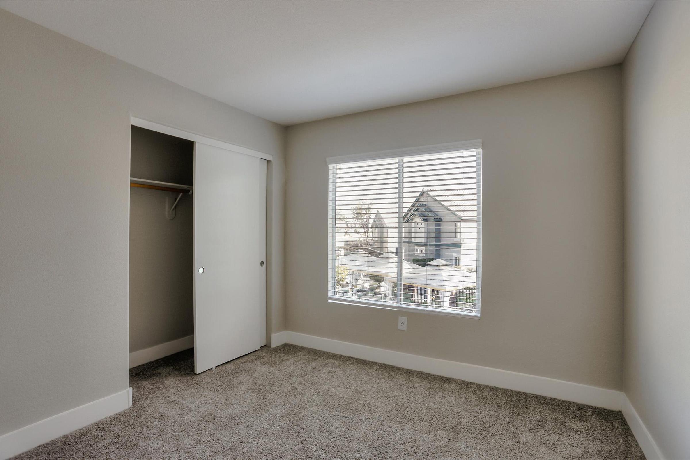 Empty room with light beige walls and grey carpeted flooring. A large window with blinds allows natural light to enter, showcasing a view of houses outside. A closet with a sliding door is present on the left side of the room. The space appears modern and minimalist.