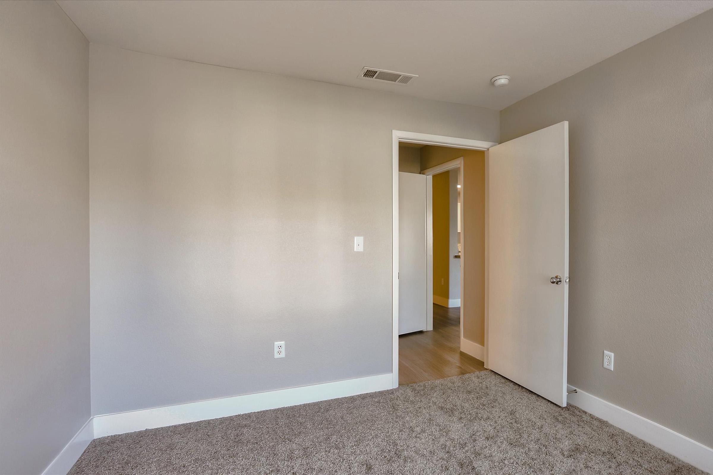 A small, empty room with light gray walls and carpeted flooring. A white door stands open, revealing another room beyond. Natural light brightens the space, emphasizing the clean and minimalist design.
