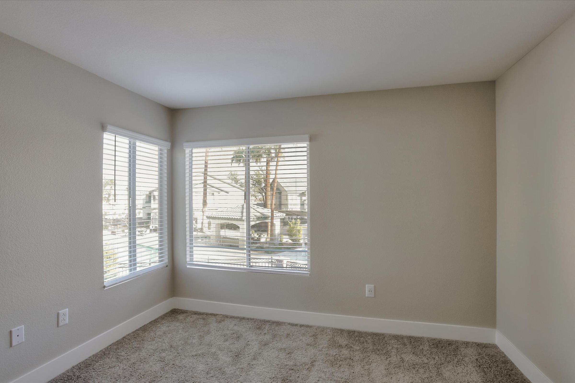 A bright, empty room with light gray walls and new carpet. Two large windows with white blinds let in natural light, offering a view of the outdoors. The room has a clean and modern aesthetic, perfect for personalizing as a living space or office.