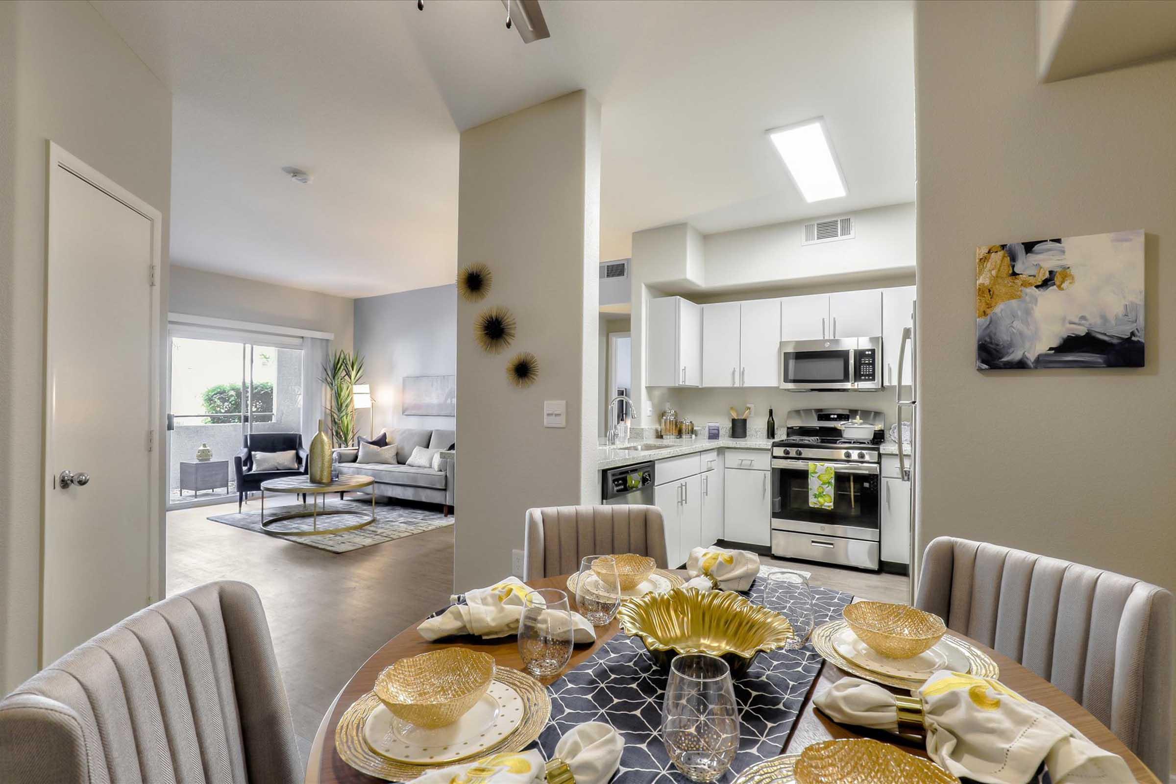 A modern dining area set for a meal, featuring a round table with elegant place settings, including gold-rimmed plates and napkins. In the background, a cozy living room is visible with a sofa, coffee table, and large windows allowing natural light, creating a warm and inviting atmosphere.