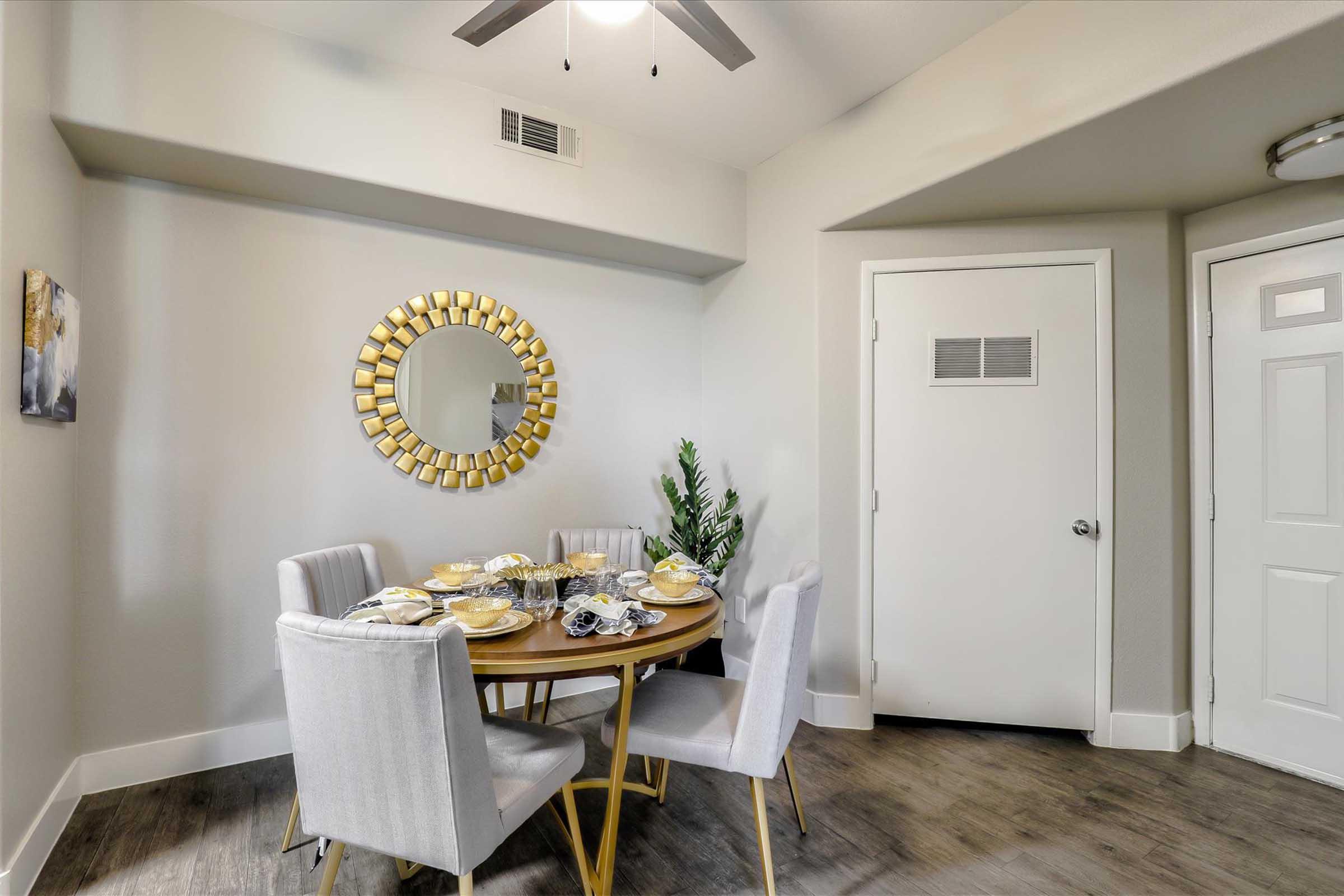 A modern dining area featuring a round wooden table set for four with elegant dinnerware. Surrounding the table are four stylish gray upholstered chairs. A sunburst mirror adorns the wall, reflecting natural light, while a green plant adds a touch of greenery. A door and light fixture are also visible.