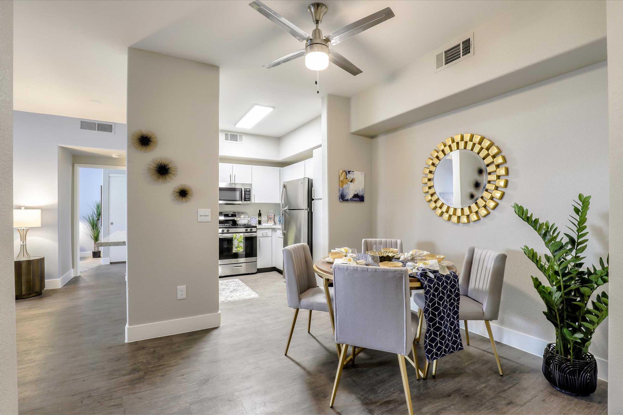A modern dining area featuring a round table set for a meal, surrounded by four upholstered chairs. A large decorative mirror is mounted on the wall, and a potted plant is nearby. The open layout includes a view of a kitchen with stainless steel appliances and a living space in the background, illuminated by soft lighting.