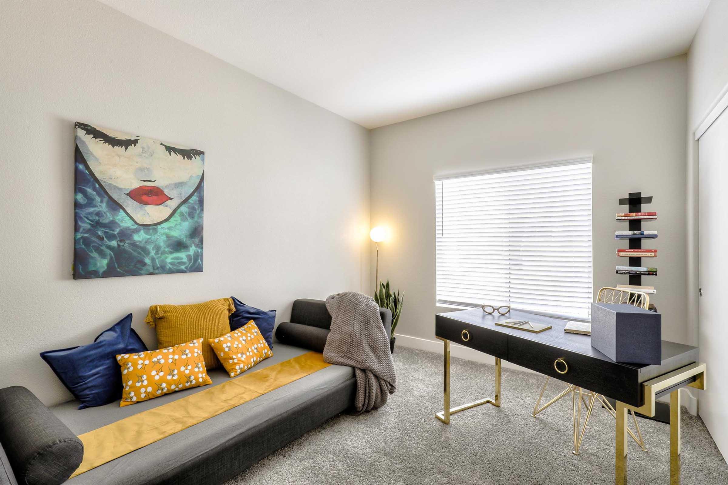 A modern, cozy room featuring a sectional sofa with colorful cushions, a stylish desk with books and a decorative item, a lamp, and a window with blinds that lets in natural light. An artwork depicting a serene face is hanging on the wall, adding a touch of creativity to the space.
