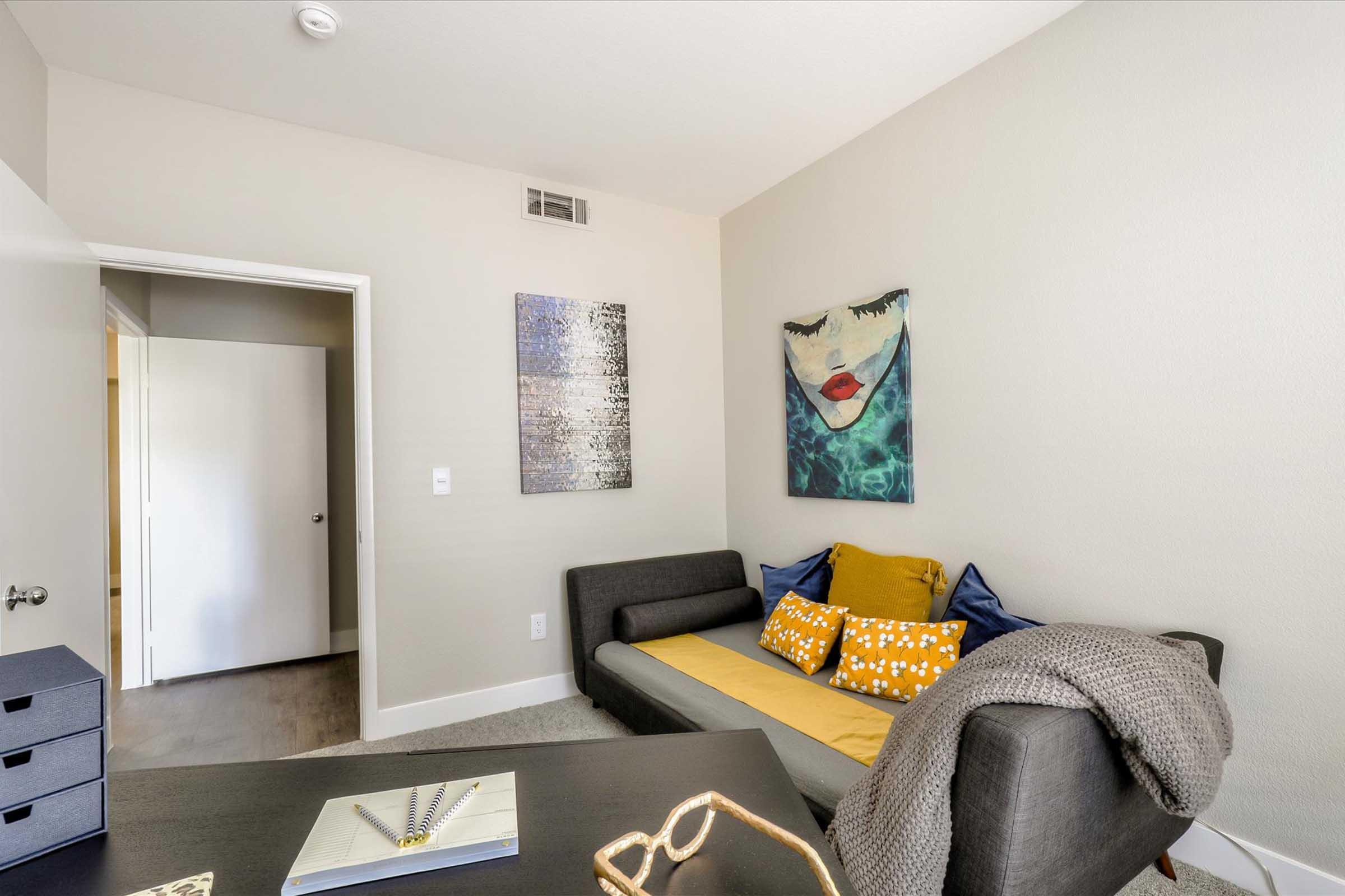A well-lit room featuring a cozy seating area with a gray sofa adorned with yellow throw pillows, a small black desk with a notebook, and a piece of abstract art on the wall. A white door leads to another room, and neutral-colored walls create a calming atmosphere.