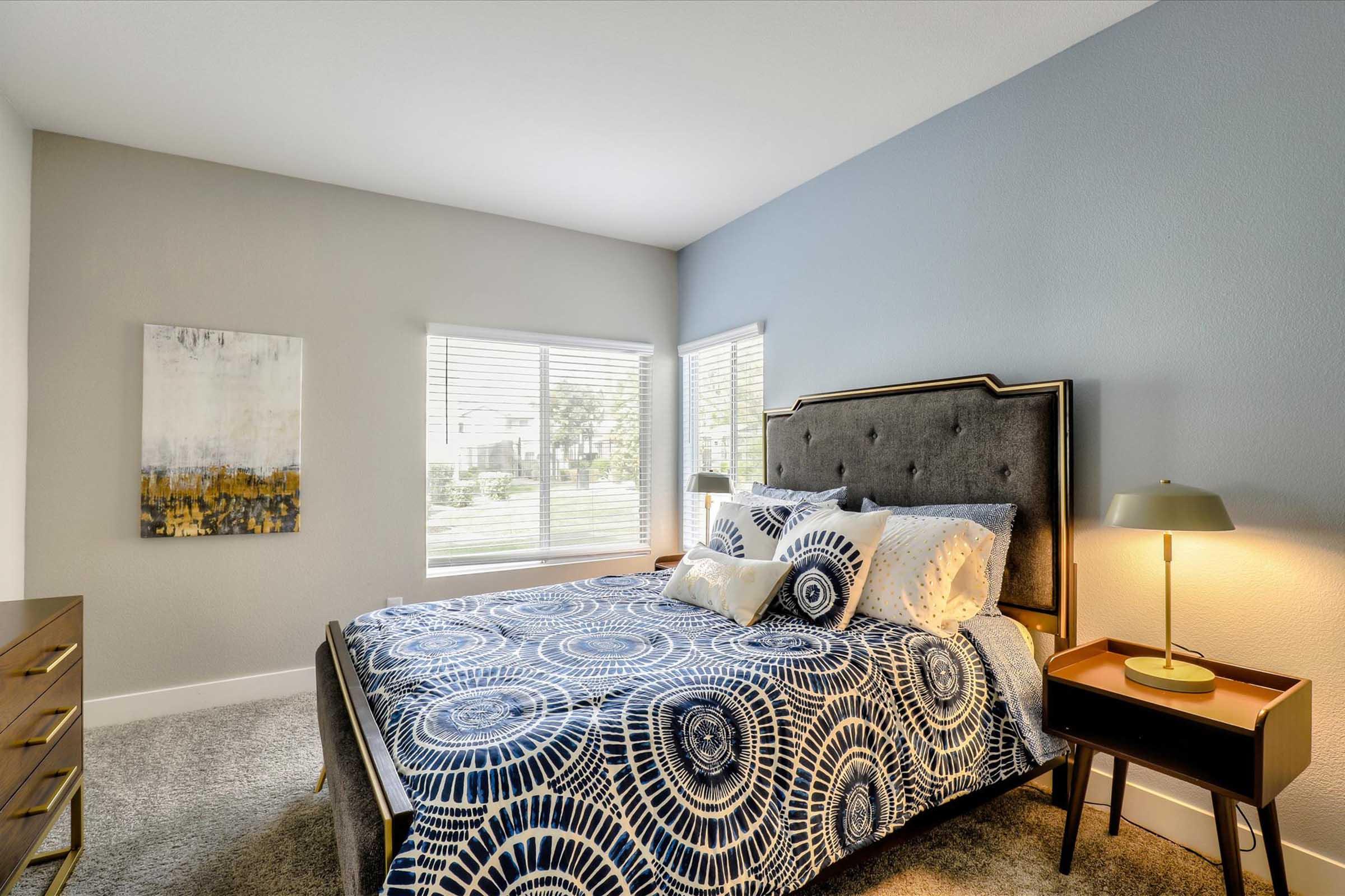 A well-lit bedroom featuring a large bed with a blue patterned duvet, decorative pillows, and a stylish headboard. There's a sleek nightstand with a lamp beside the bed. A modern dresser is visible, and the room has two windows with blinds, allowing natural light to enter. A piece of abstract art hangs on the wall.
