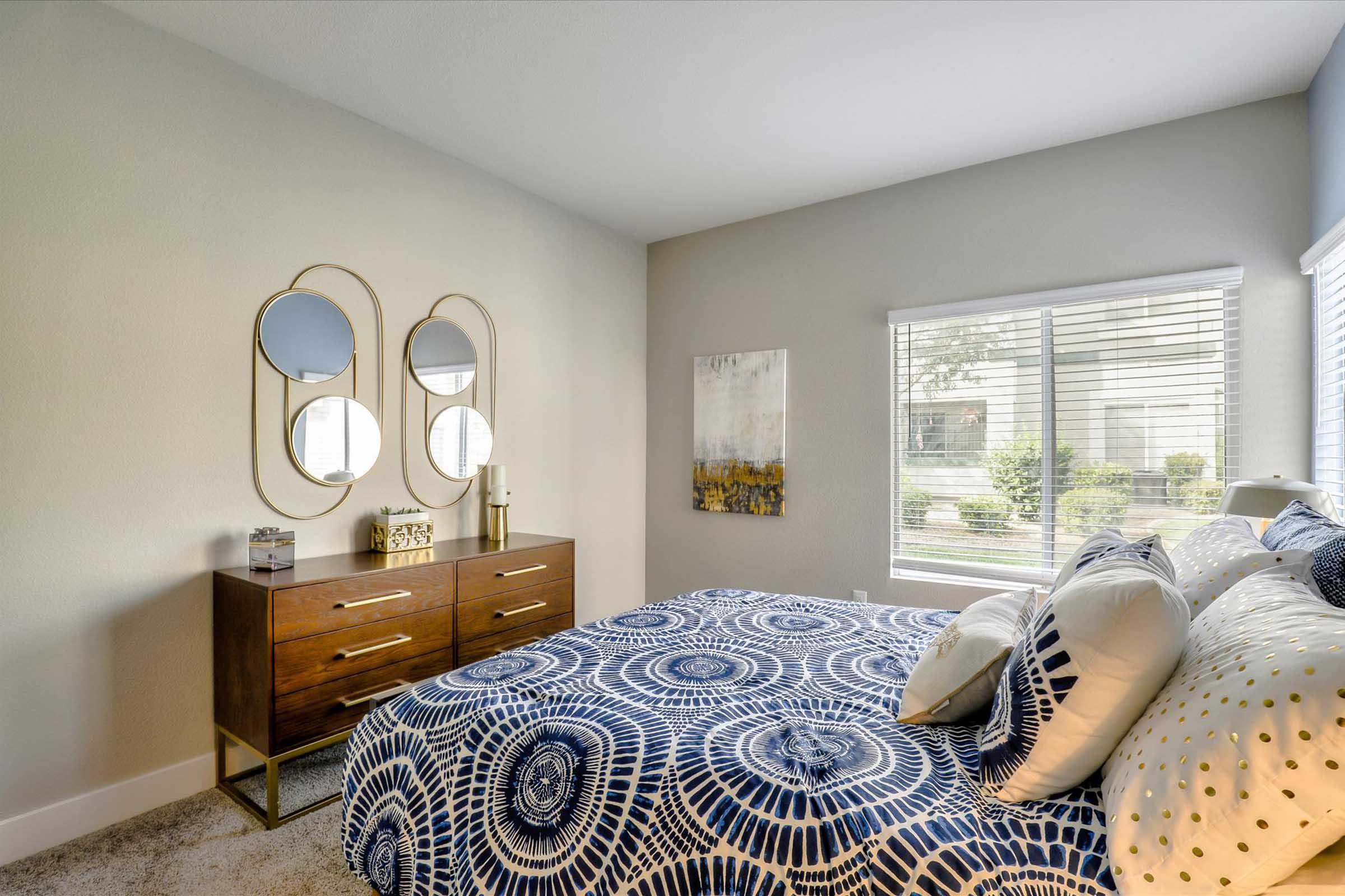A cozy bedroom featuring a bed with a blue and white patterned duvet, complemented by decorative pillows. A modern wooden dresser with circular mirrors hangs above it. The room has large windows allowing natural light, and there's artwork on the wall, creating a stylish and inviting atmosphere.