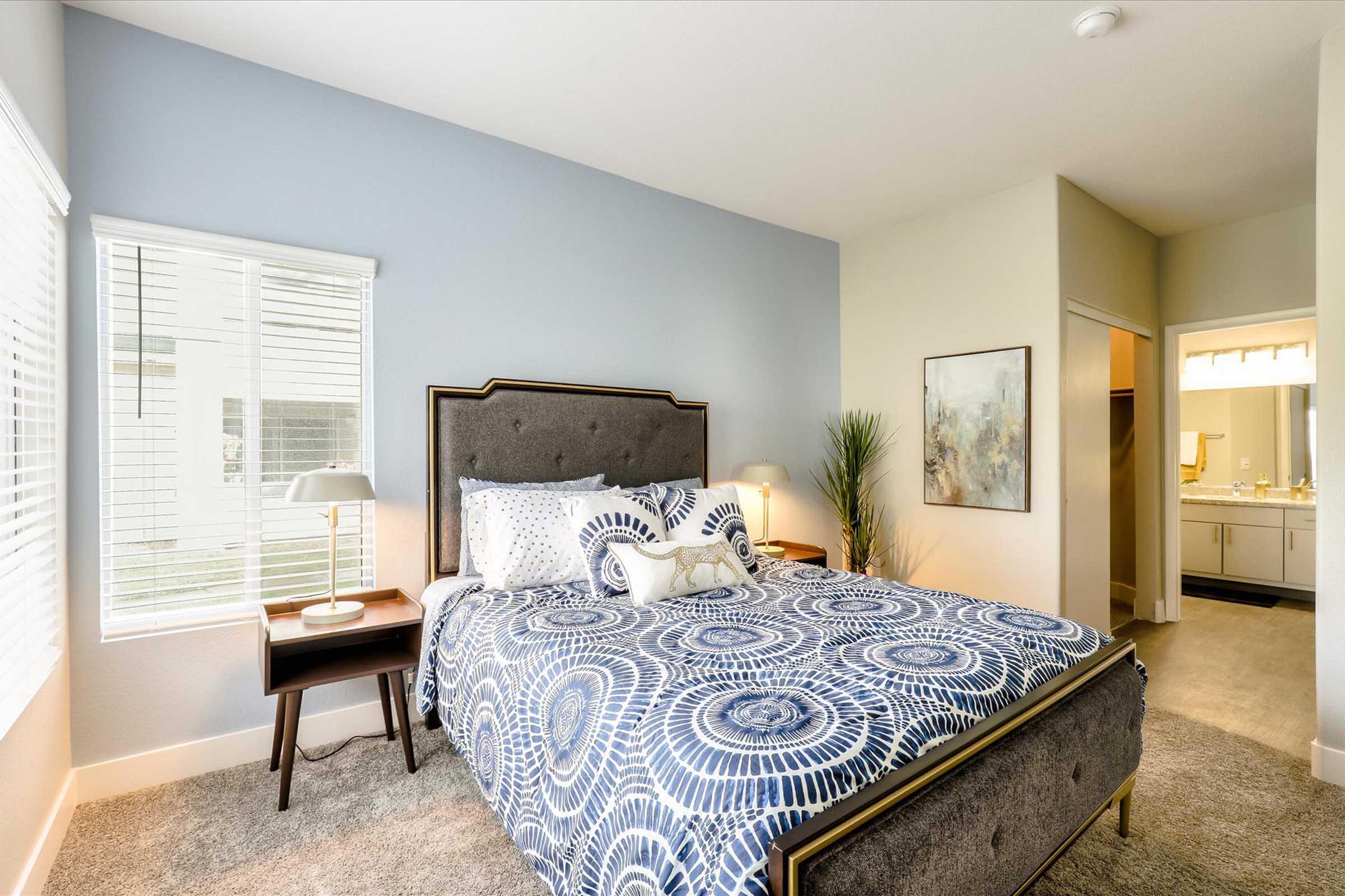 Cozy bedroom featuring a queen-sized bed with a patterned blue and white duvet, a bedside table with a lamp, and a large window letting in natural light. Neutral-colored walls and carpeting create a warm atmosphere, with a doorway leading to a bathroom. A plant adds a touch of greenery to the room.