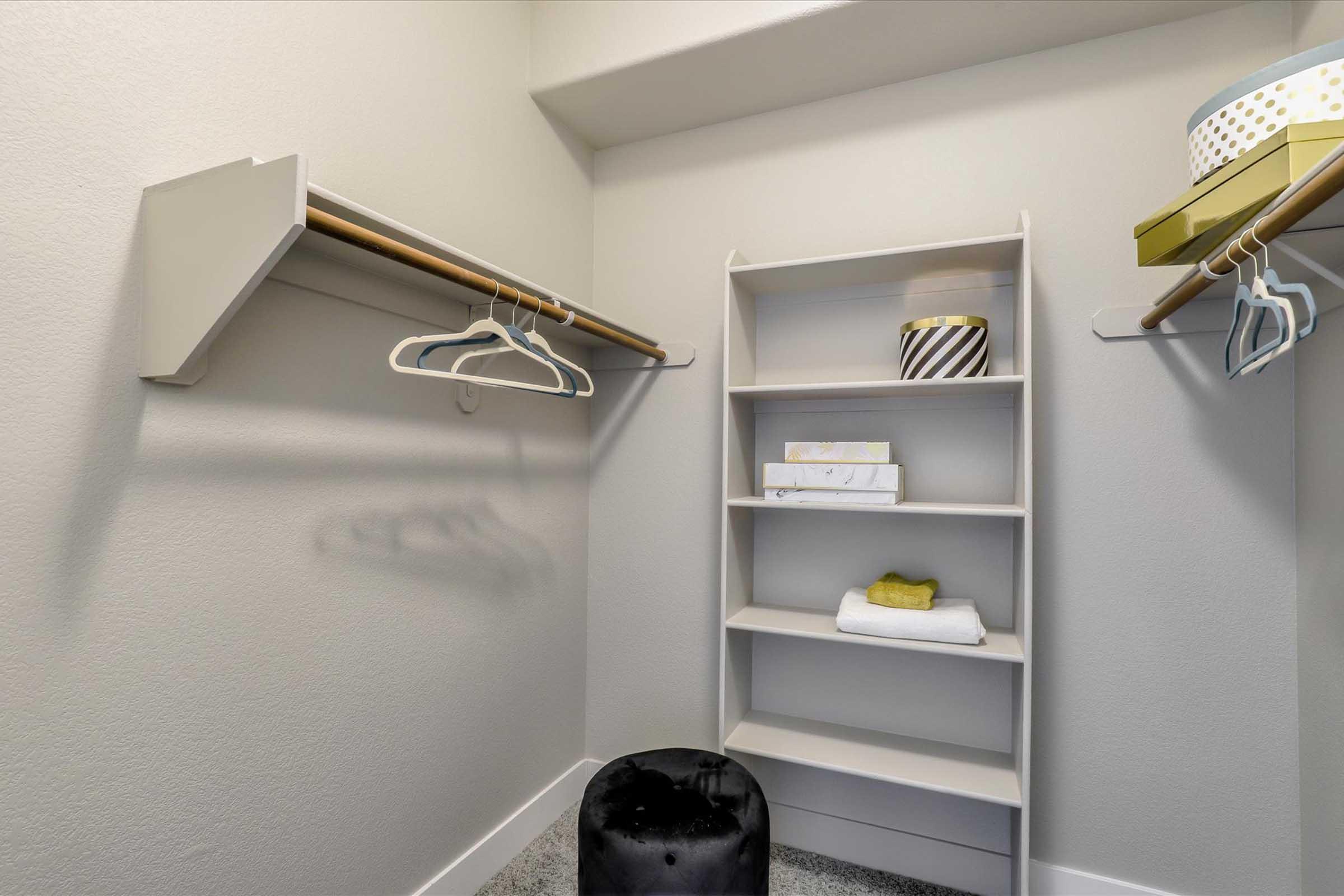 A spacious closet featuring two hanging rods, one on the left and one on the right, with several white hangers. In the center, there’s a light-colored shelf unit holding neatly folded towels and decorative boxes, along with a small black ottoman on the floor. The walls are a soft gray.
