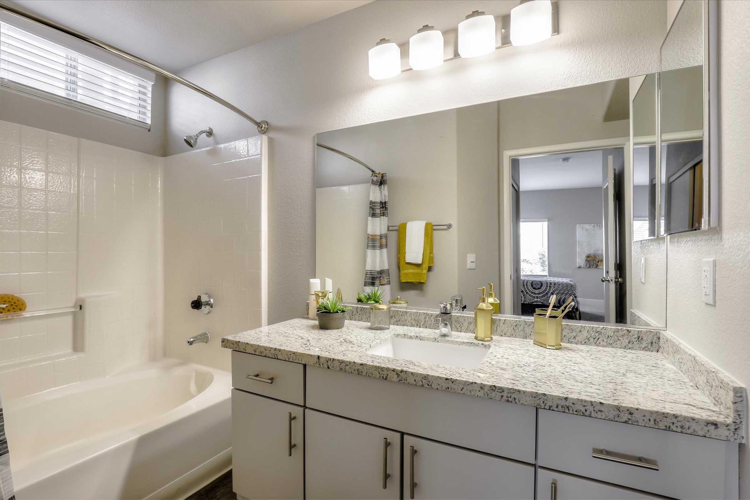 A modern bathroom featuring a combined bathtub and shower, a spacious double vanity with granite countertop, two sinks, and a large mirror. Natural light enters through a window, and towels are neatly hung on a nearby rack. Decorative plants add a touch of greenery to the space.