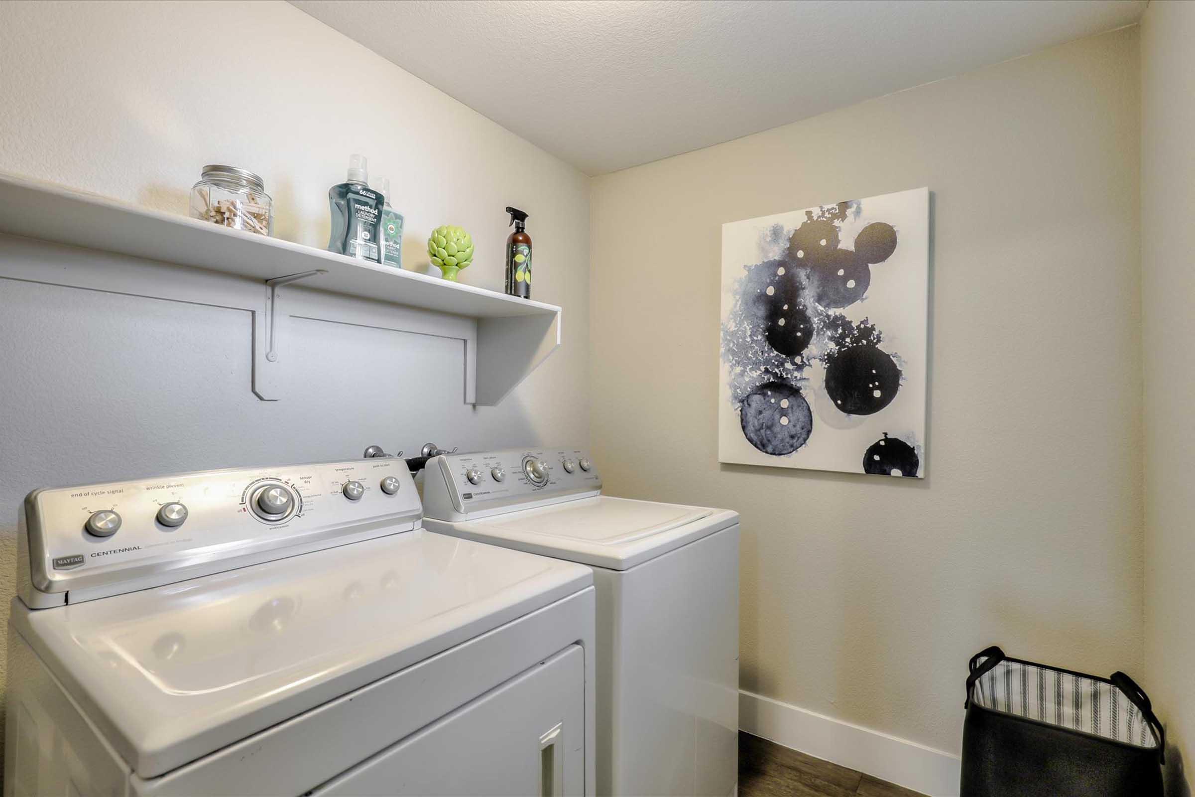 A laundry room featuring two white washing machines, a shelf with decorative items, and a wall art piece with abstract blue bubbles against a light background. A black and white striped laundry basket is positioned in the corner. The space is well-lit with neutral-colored walls and a clean, organized appearance.