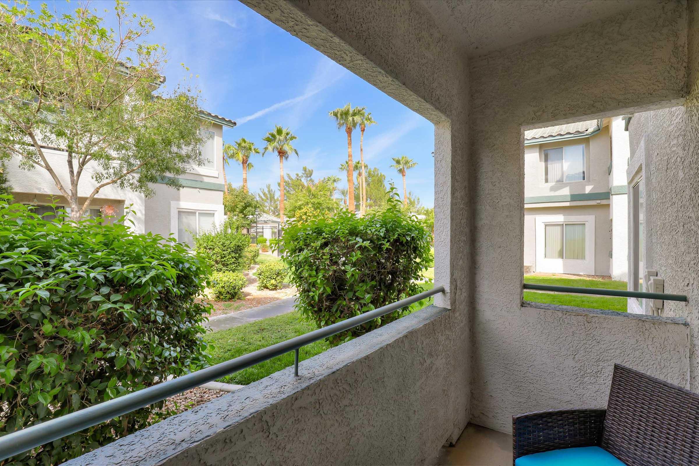 A view from a balcony overlooking a landscaped courtyard with shrubs and palm trees. The scene includes two apartment buildings in the background under a clear blue sky. A comfortable chair is visible on the balcony railing.
