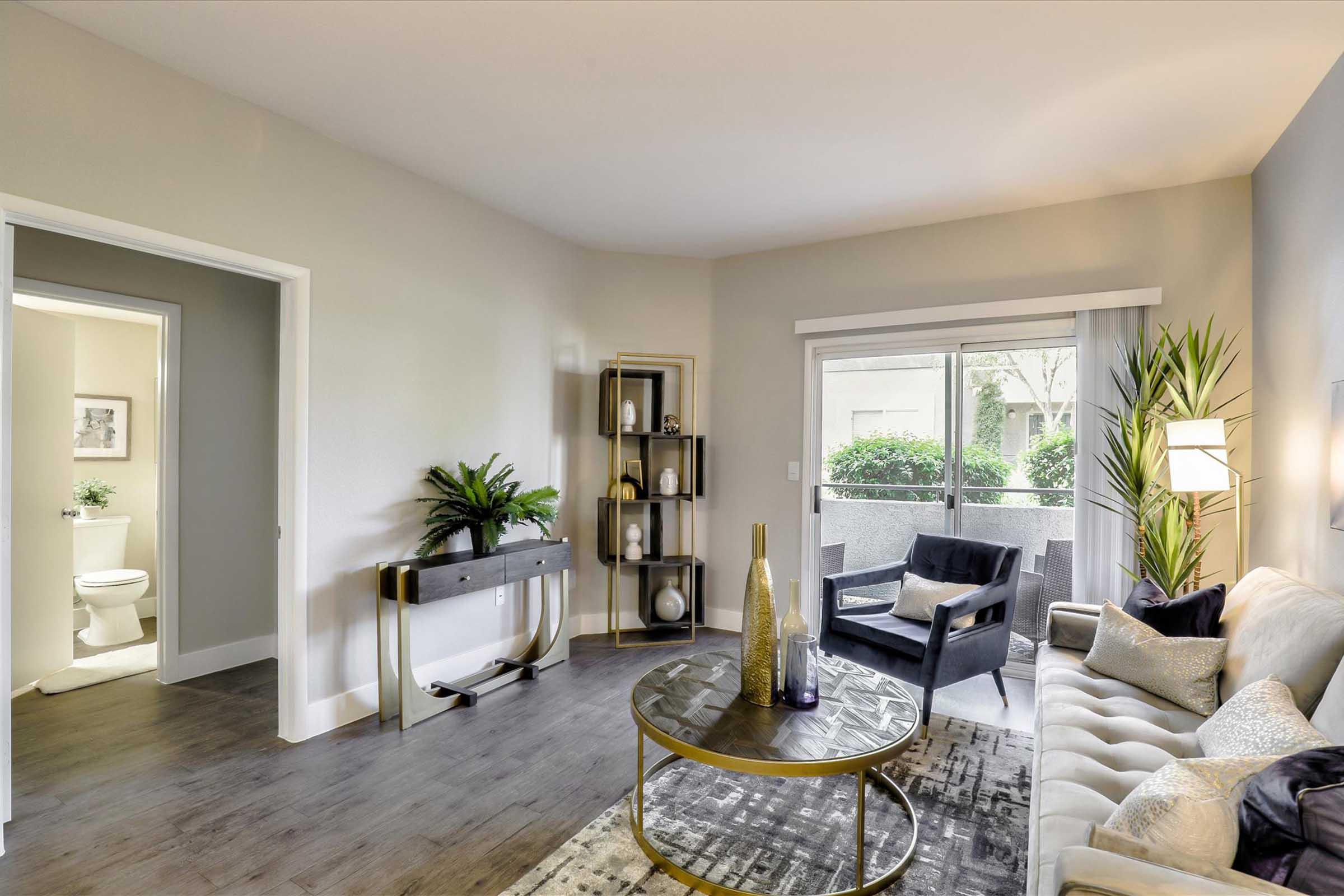 A modern living room featuring a light-colored sofa with decorative pillows, a stylish round coffee table, and a console table with plants. A large window provides natural light, and there's an armchair next to it. A door leads to a bathroom, adding to the room's contemporary decor.