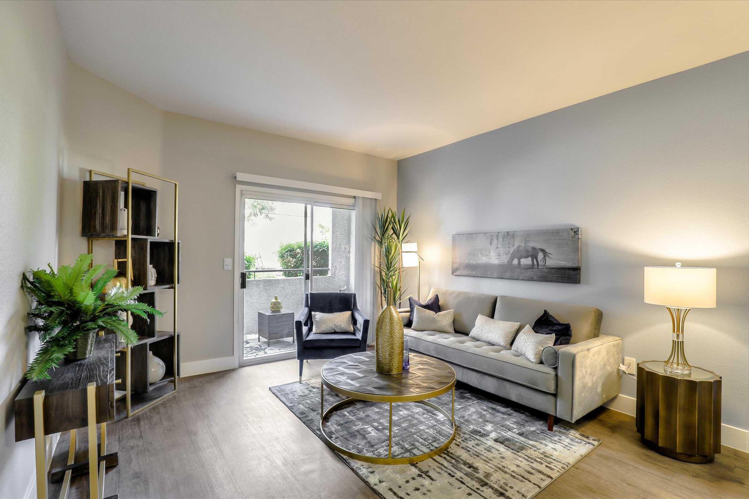 A stylish living room featuring a light gray sofa with decorative pillows, two dark chairs, a round coffee table, and a floor lamp. A potted plant and a shelf with decorative items add to the decor. Natural light streams through a window with a view of greenery outside, enhancing the inviting atmosphere.