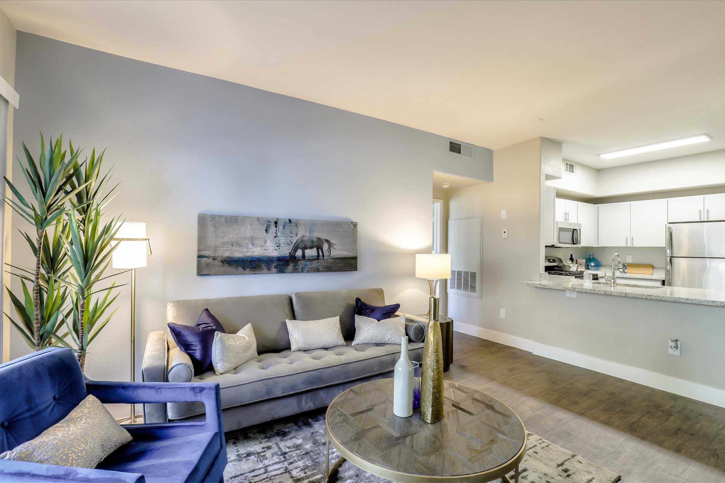 A modern living room featuring a gray sofa with decorative pillows, a circular glass coffee table, a potted plant, a floor lamp, and a wall art piece depicting a horse. In the background, a well-equipped kitchen with stainless steel appliances is visible.