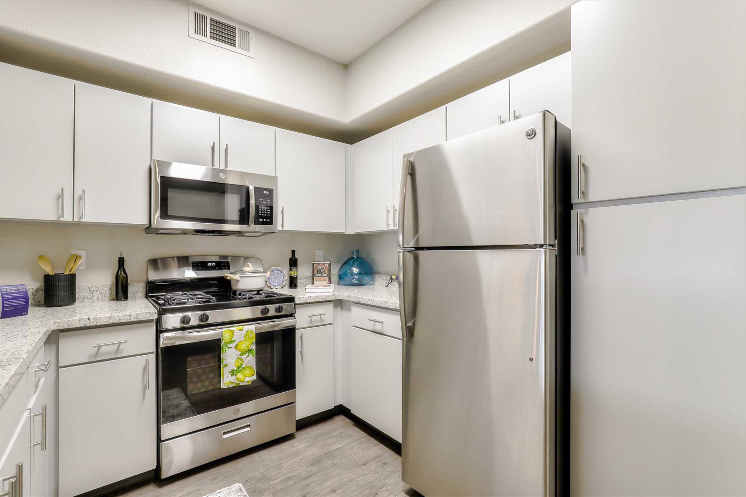 A modern kitchen featuring white cabinets, stainless steel appliances including a microwave and refrigerator, a gas stove, and a light granite countertop. Decor items include a blue glass vase and kitchen utensils, with a towel hanging from the stove for a touch of color.