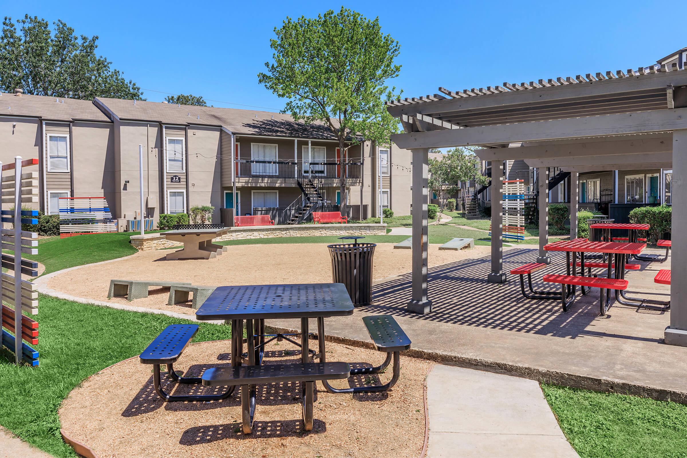 A well-maintained outdoor area featuring a picnic table, benches, and play equipment surrounded by green lawns. The scene includes a sand area and nearby buildings, providing a welcoming space for social gatherings and recreation under clear blue skies.