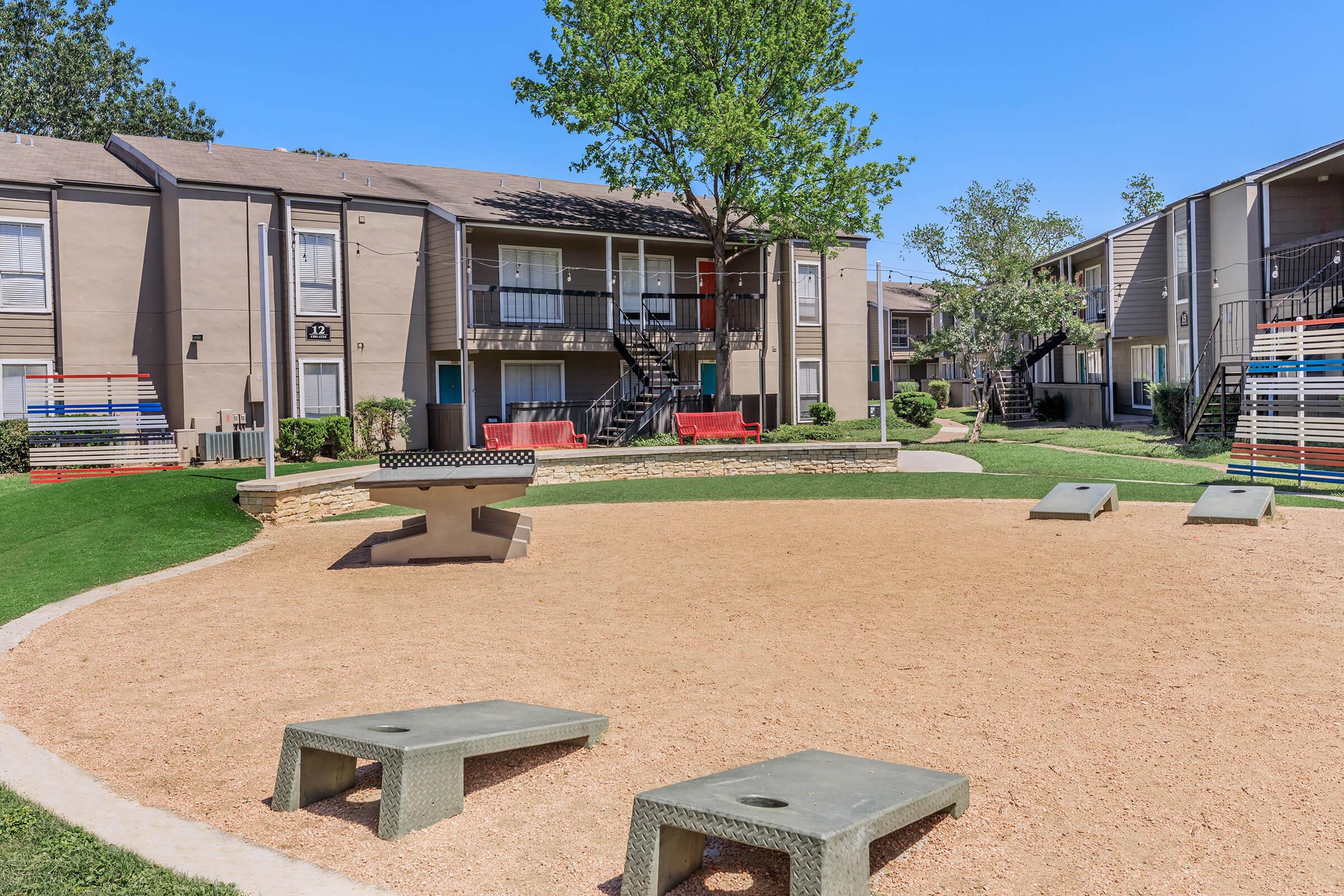 A well-maintained outdoor apartment complex featuring a sandy area with two stone-style seating benches, surrounded by lush green grass. In the background, there are two buildings with balconies and colorful seating areas. The sky is clear and blue, creating a bright and inviting atmosphere.