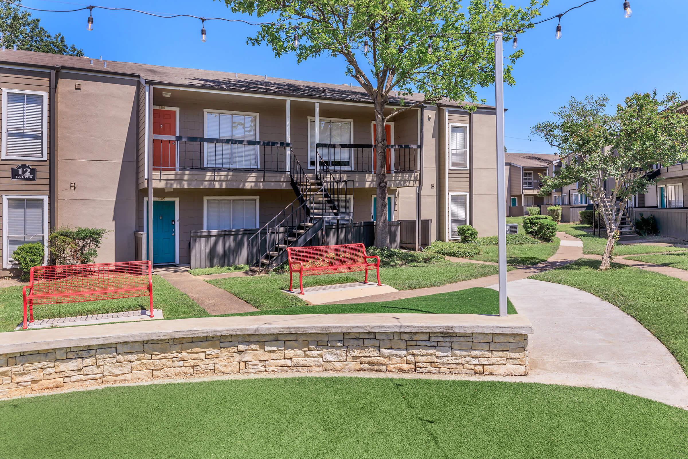 A well-maintained apartment complex featuring two-story buildings, green lawns, and walking paths. The scene includes red benches and string lights, providing a welcoming outdoor environment. Lush trees and shrubs add greenery to the surroundings, creating a pleasant living space.