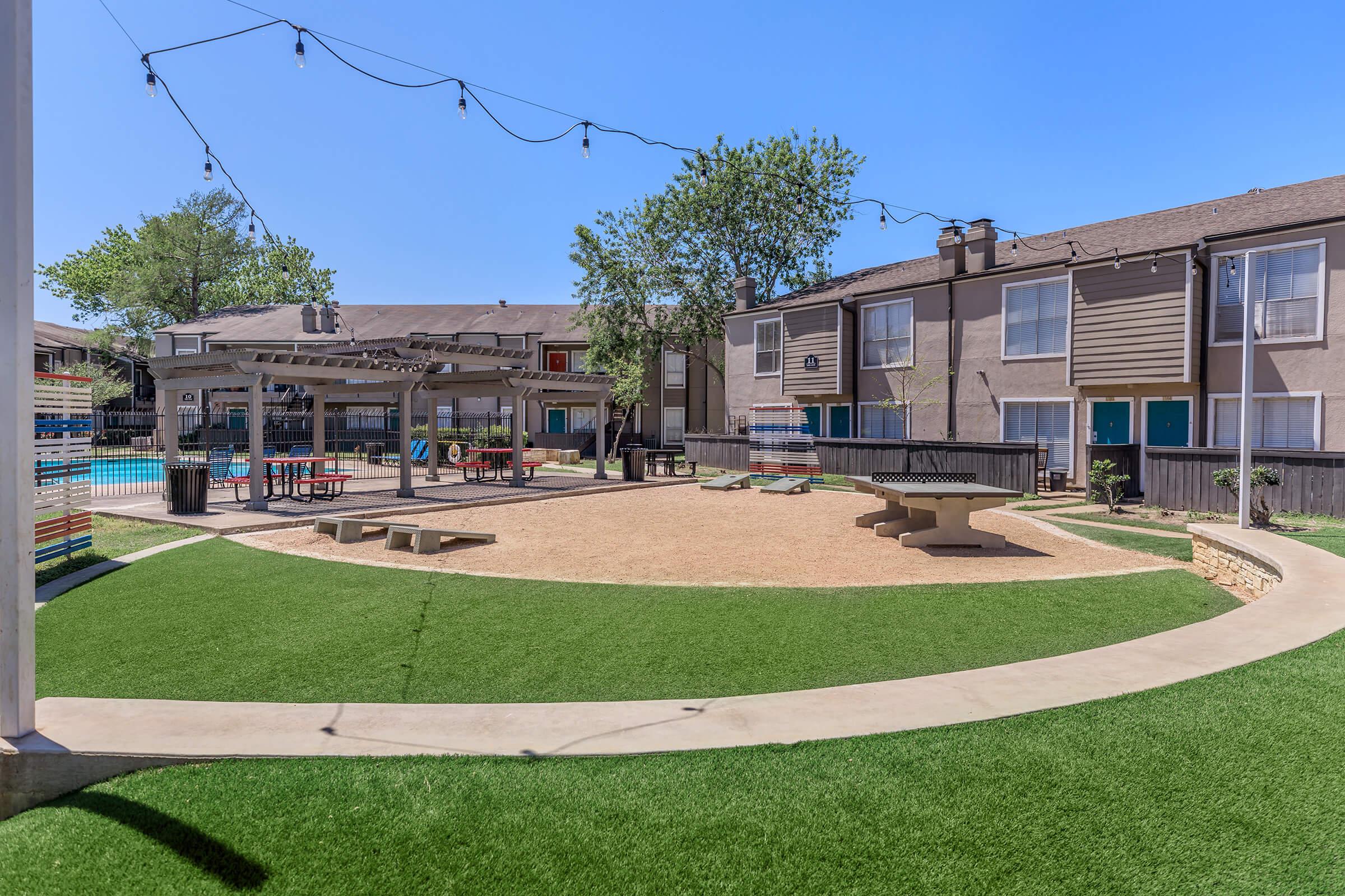 A spacious outdoor area featuring a well-maintained lawn, a sandy fire pit area, and a shaded pavilion with seating. Surrounding the space are residential buildings, with a swimming pool visible in the background and string lights overhead, adding a cozy ambiance.