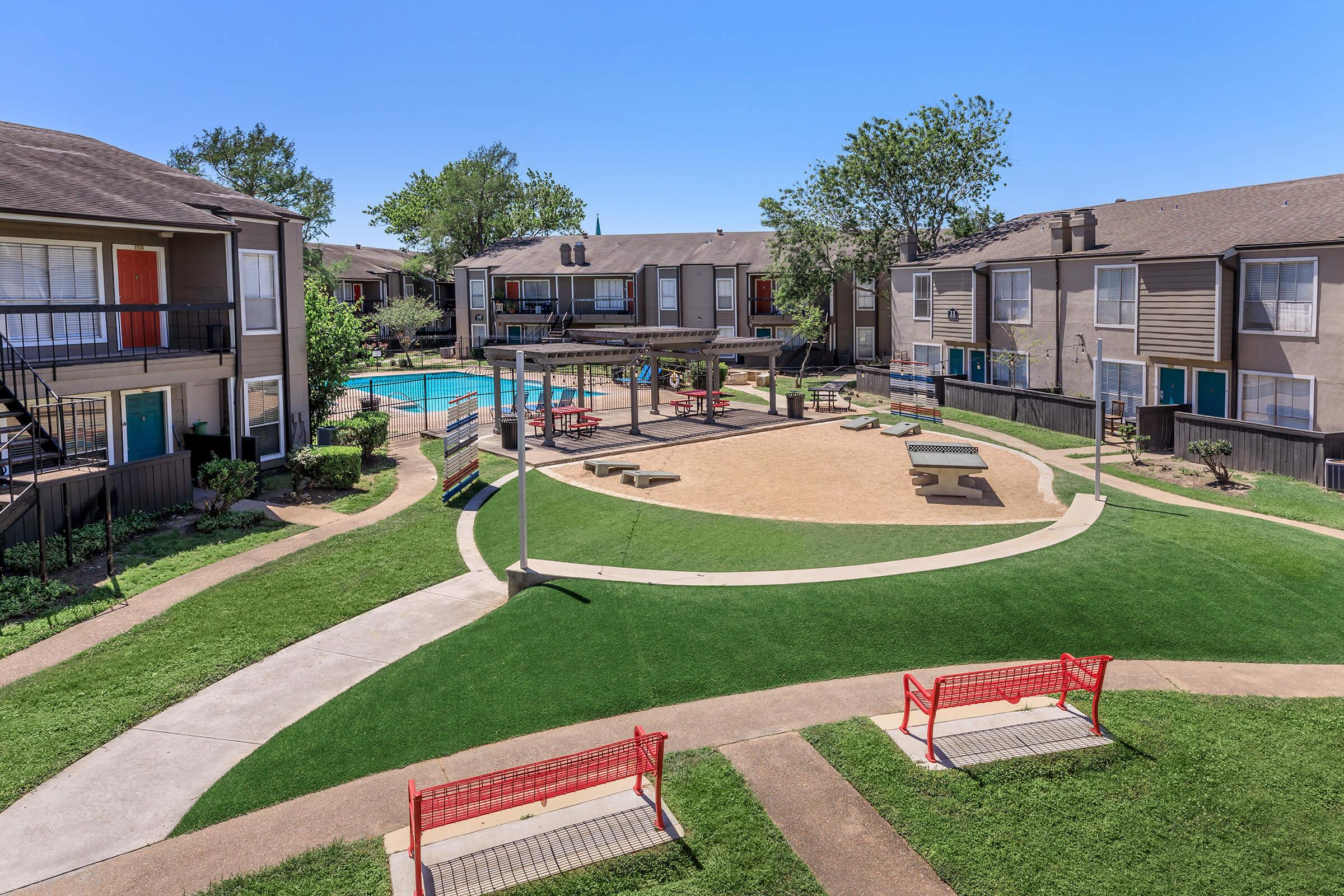 A spacious apartment complex courtyard featuring a sandy play area, picnic tables, and a swimming pool. Lush green lawns surround the area, with red benches and pathways leading to the buildings. Bright, clear skies enhance the inviting atmosphere of the community space.