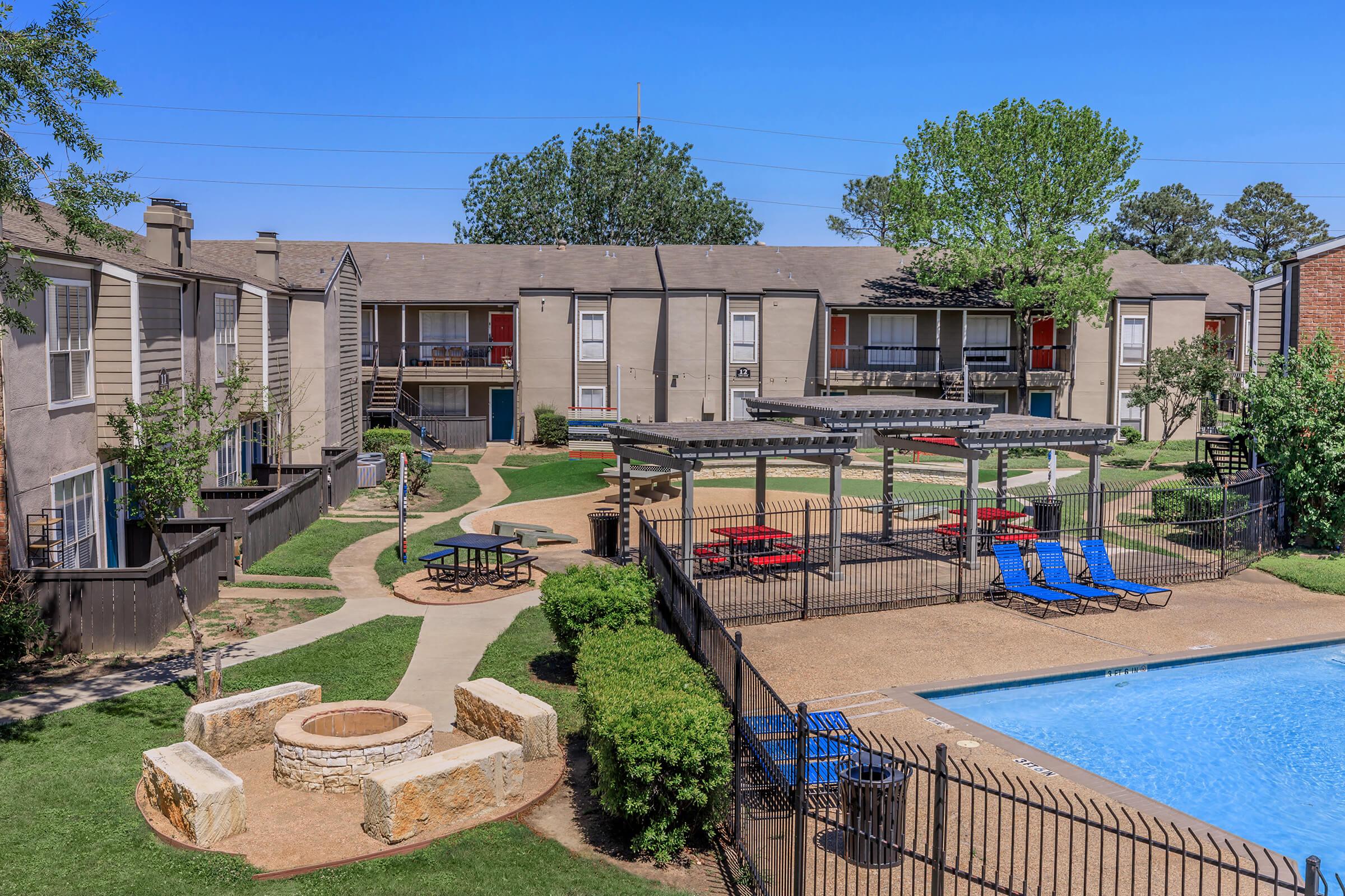 A scenic view of an apartment complex featuring a swimming pool, surrounded by well-maintained landscaping. In the foreground, there is a fire pit area with stone seating, and picnic tables nearby. The complex includes multiple buildings with balconies and green trees in the background under a clear blue sky.