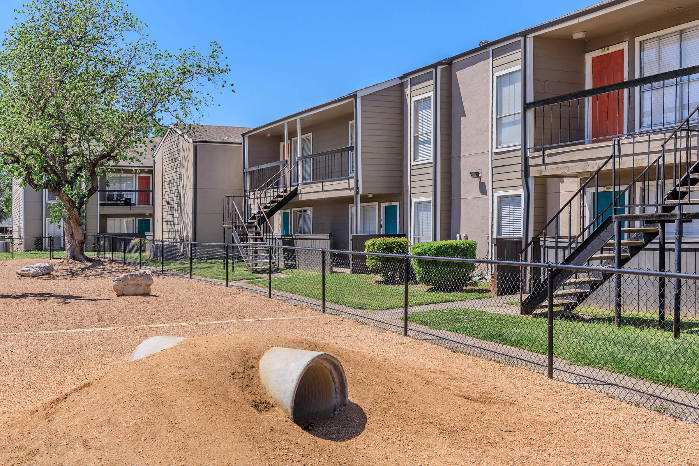 Residential apartment complex with two-story buildings. Features a fenced sandy area with a concrete pipe in the foreground and well-maintained landscaping. Several balconies visible, and a large tree provides shade. Clear blue sky overhead enhances the vibrant setting.