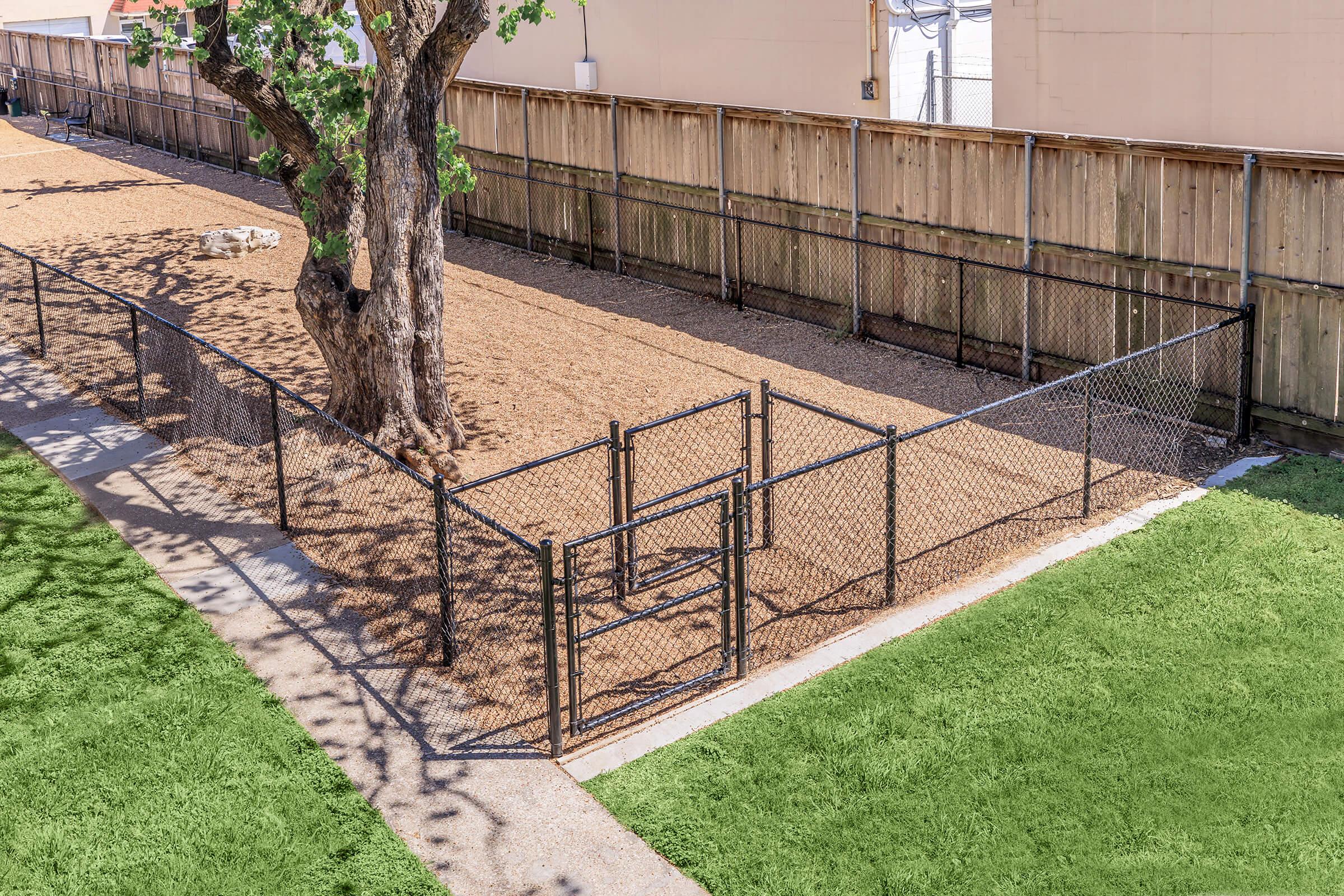 A fenced dog park featuring a sandy area surrounded by a chain-link fence. In the center, there is a gated entrance. A large tree stands nearby, providing some shade. The park is bordered by a wooden fence, with pathways on either side, and grass areas visible in the foreground.