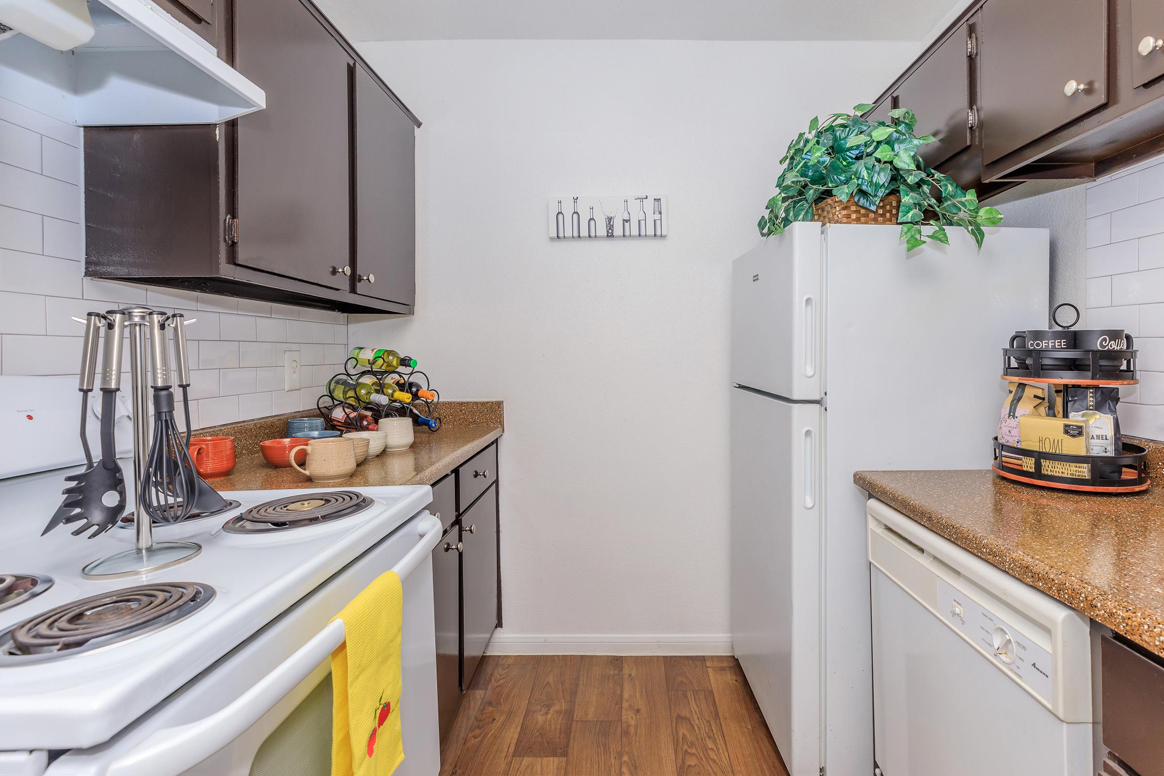 A modern kitchen featuring dark wooden cabinets and a light-colored countertop. There’s a stove, a refrigerator, and a dishwasher. Countertop items include fruit in a bowl, mugs, and cooking utensils. A decorative plant is visible on top of the cabinets, and there are wall decorations with kitchen-themed artwork.