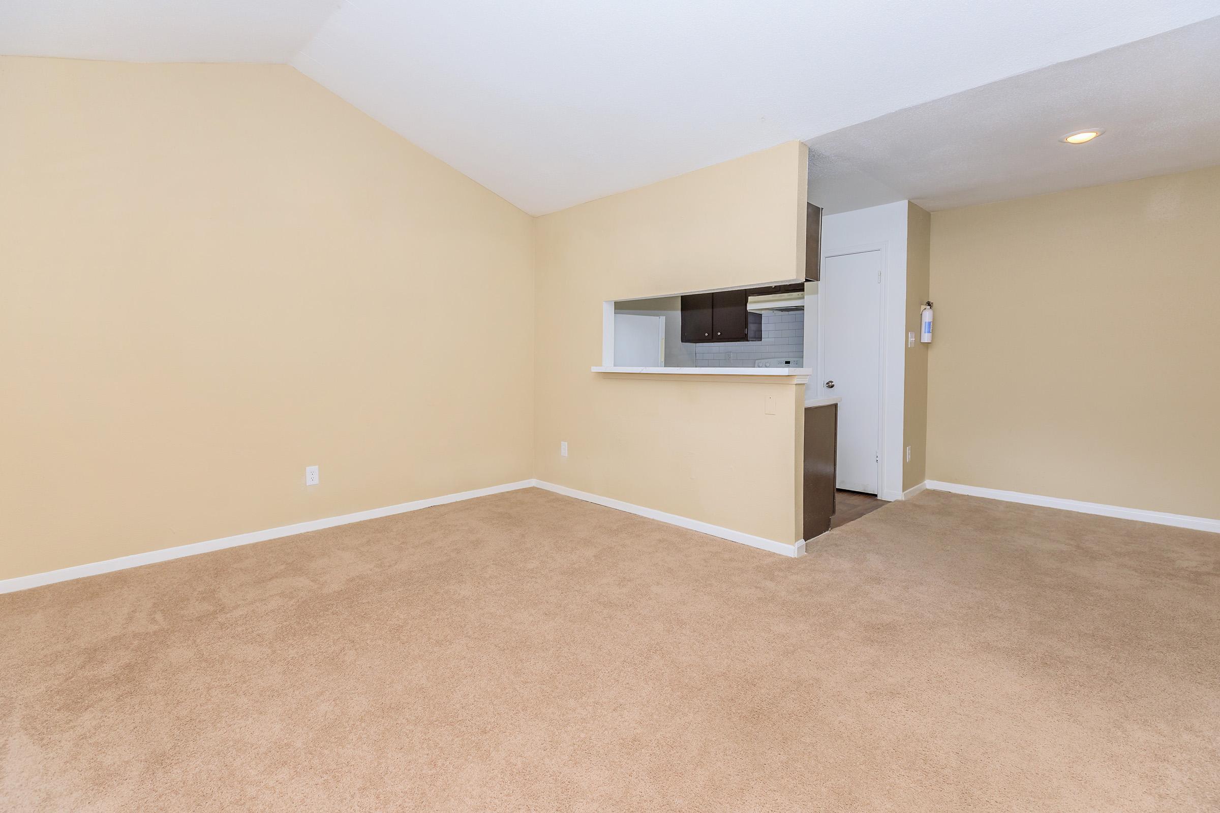 Spacious, empty indoor area featuring beige walls and carpet, with a small kitchen counter visible in the background. A doorway leads to another room, and the ceiling is slightly vaulted, creating an airy feel. Natural light is present, enhancing the openness of the space.