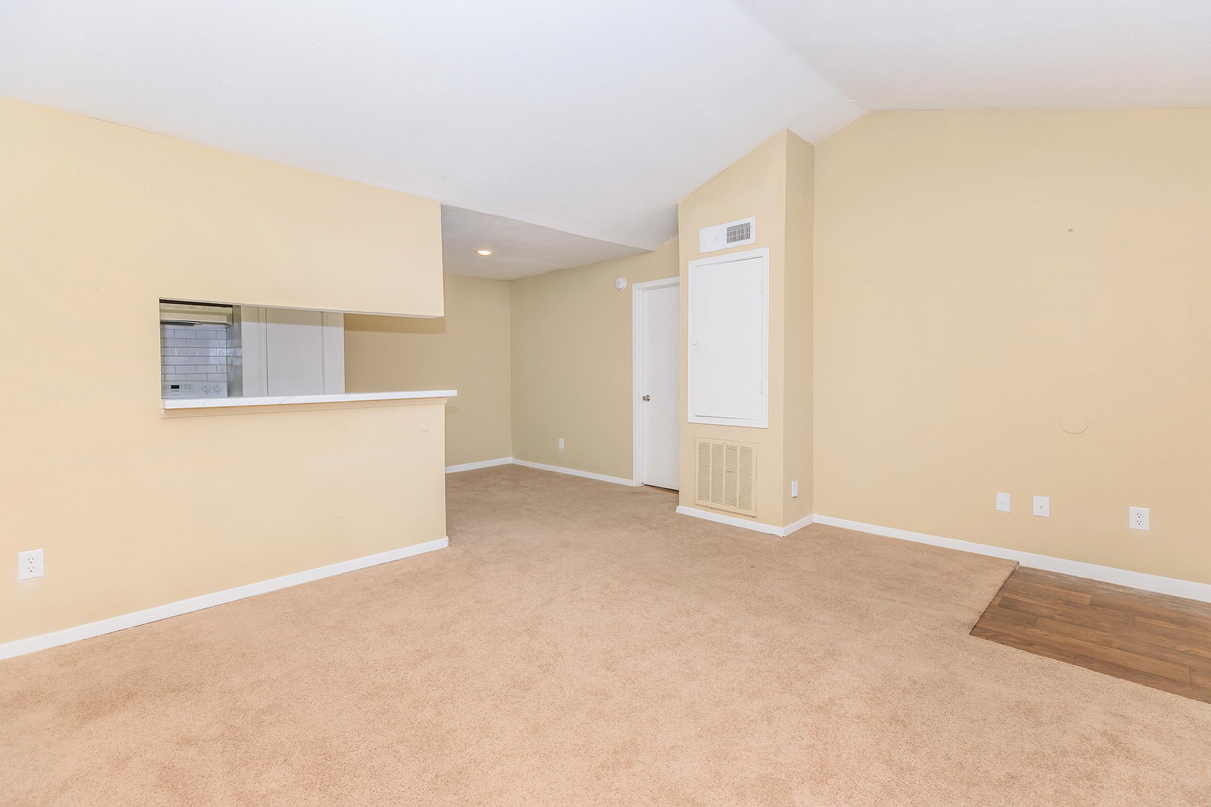 Empty living room with tan carpet and light beige walls. There is a small alcove with a countertop that opens to another room. A white door leads to a separate space, and a small vent is visible on the wall. The room is well-lit with natural light coming from a window out of view.