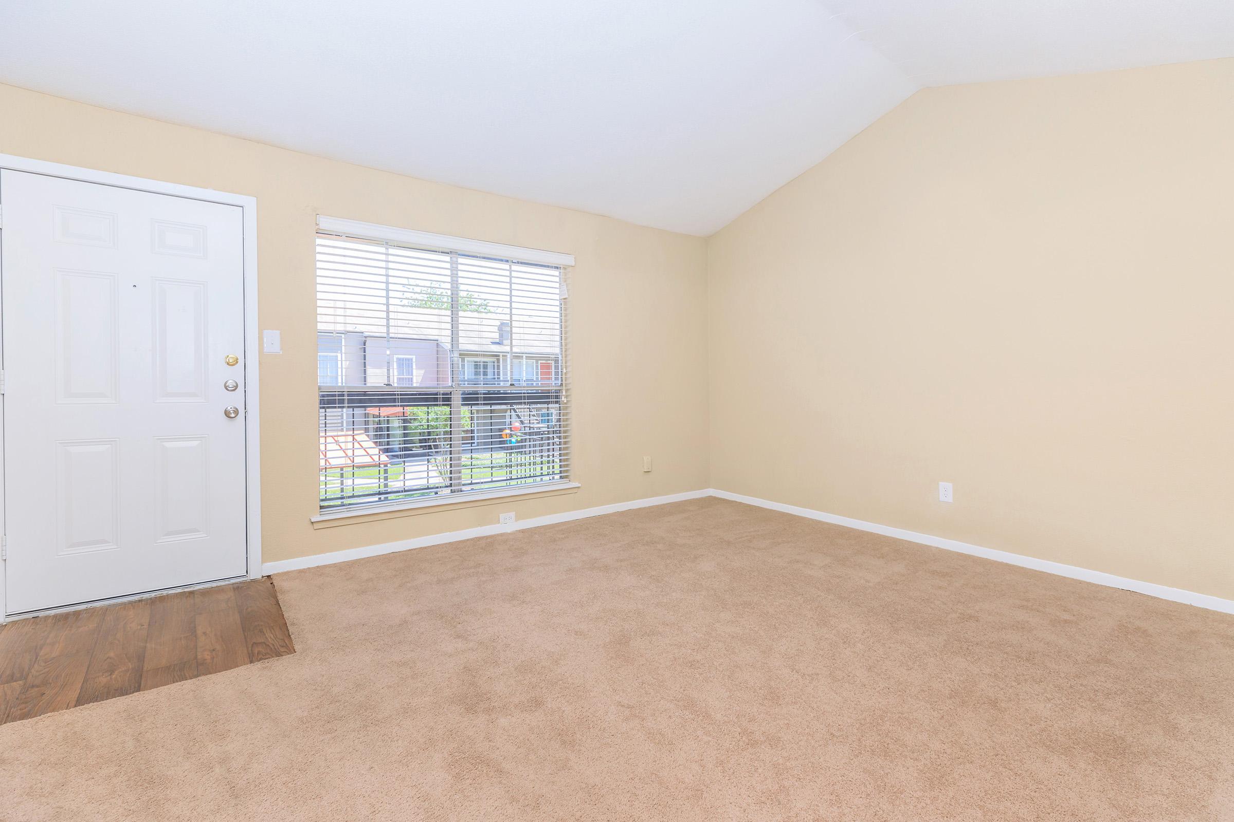 A spacious, empty living room featuring light beige walls, a large window with blinds allowing natural light, and a door leading outside. The floor has a combination of carpet and wood finish, creating a cozy atmosphere. The room has high ceilings, enhancing its openness.