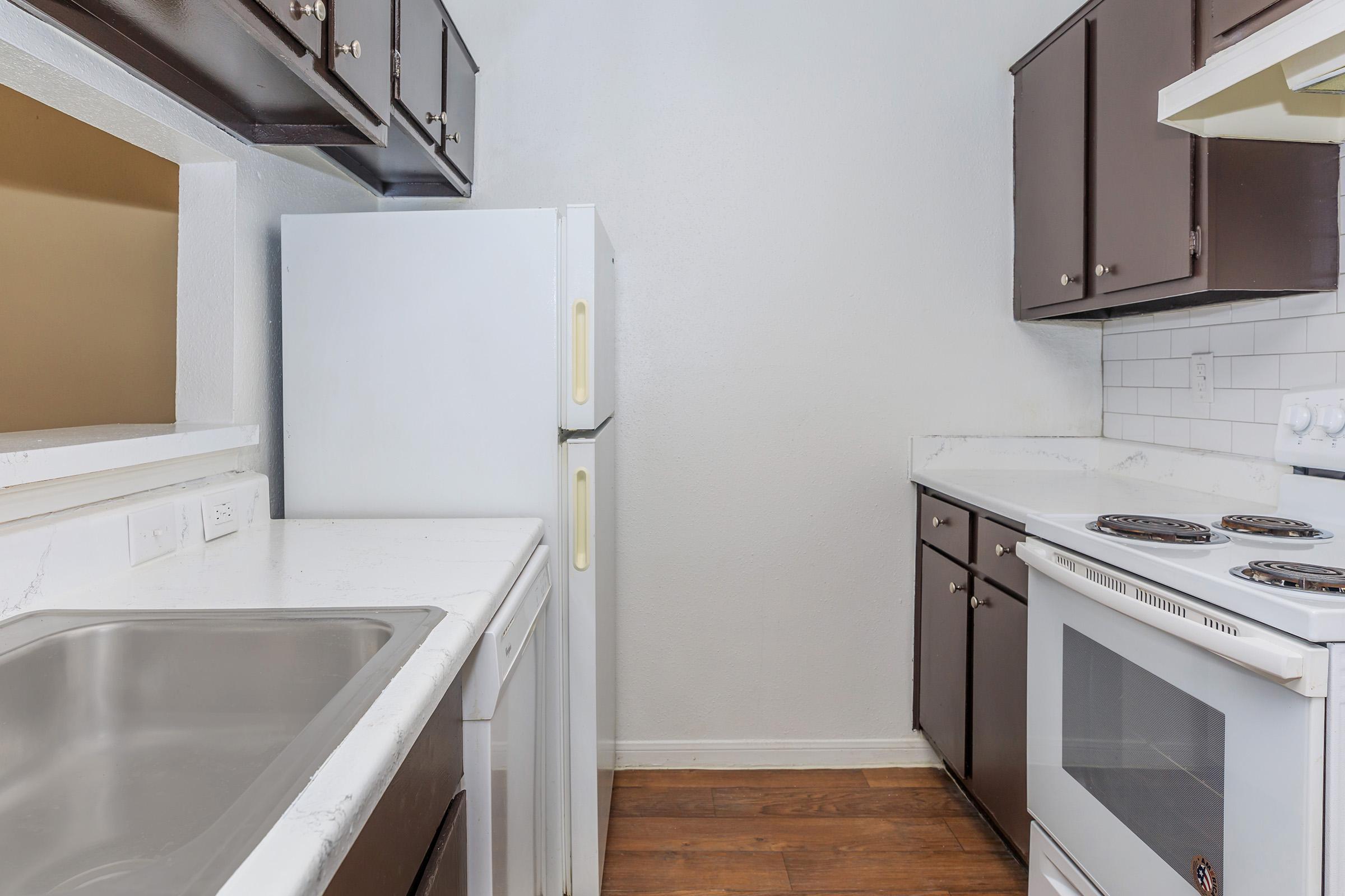 A small, modern kitchen featuring white countertops, a stainless steel sink, and dark wood cabinetry. A white refrigerator is positioned against one wall, while the opposite wall has a stove and oven. The flooring is a warm wood finish, creating a cozy atmosphere.