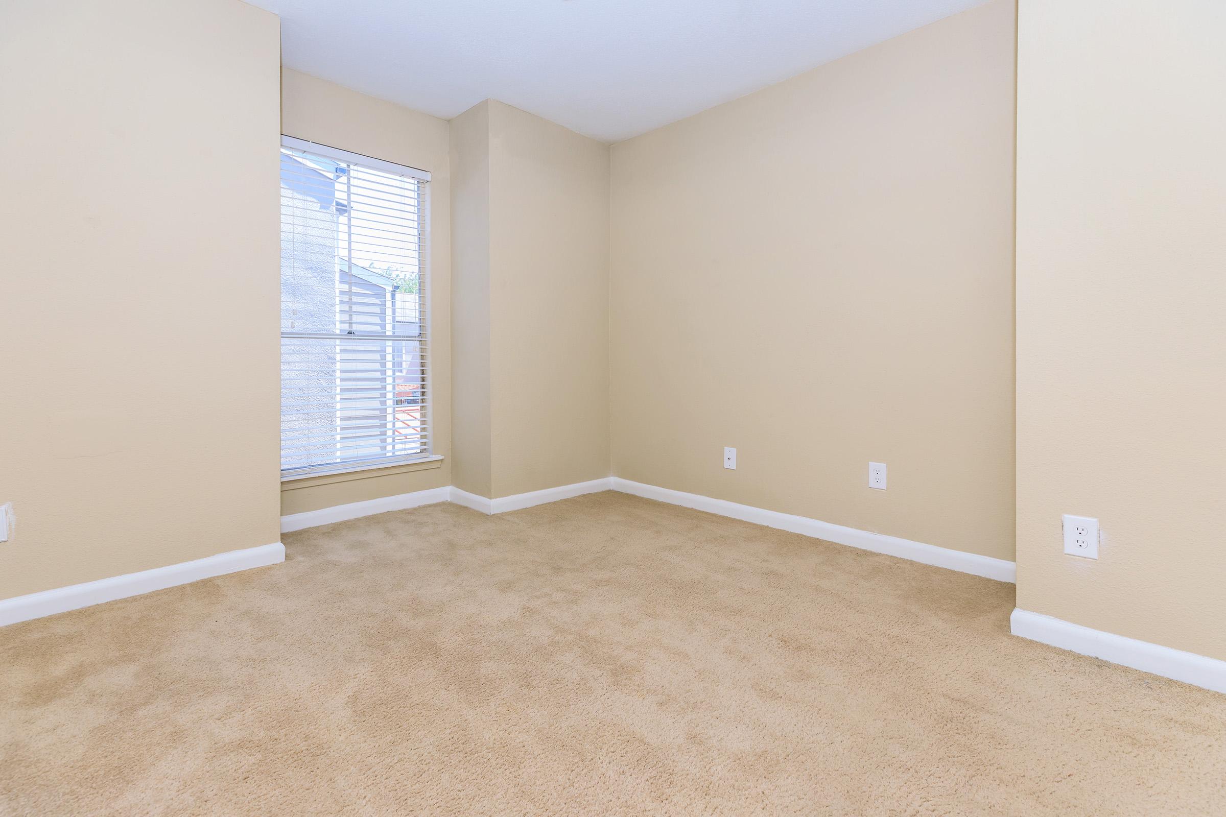 A small, empty room with light beige walls and carpet. A window with white blinds on one wall allows natural light to enter, while the opposite wall is bare. The corner of the room creates a cozy, intimate feel, perfect for personal decoration or furniture arrangement.