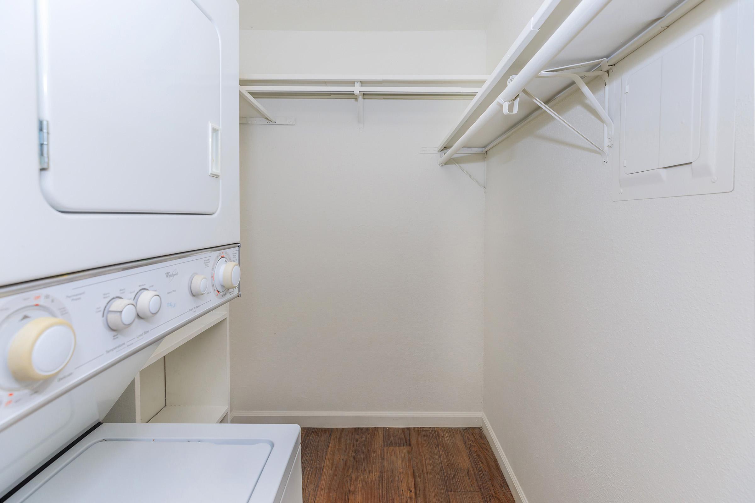 A small laundry room featuring a stacked washer and dryer against a light wall. There's a white shelving unit above for storage, and the floor is made of wood laminate. The space is uncluttered and bright, with plenty of open wall space visible.