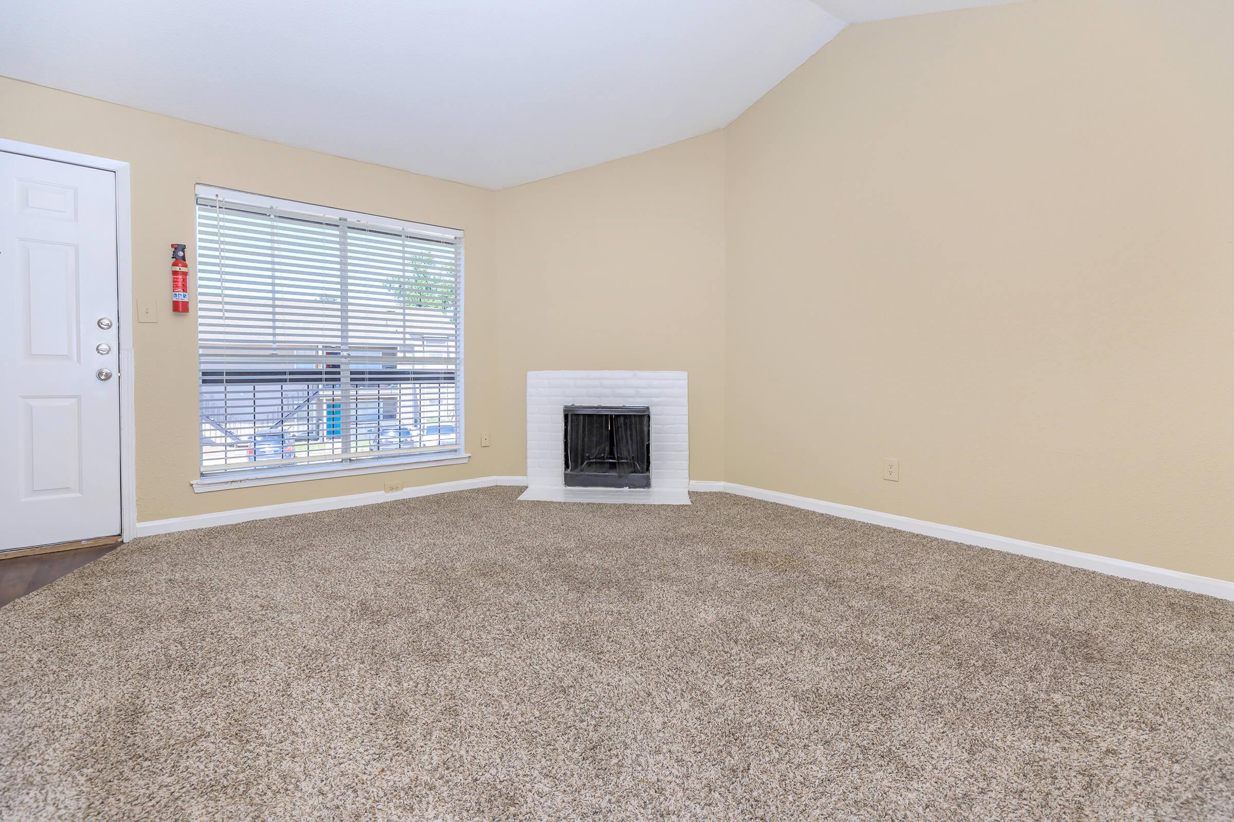 A spacious, empty living room featuring light beige walls, a large window with blinds, and a white, brick fireplace. The floor is covered in plush, light brown carpet. The entrance door is visible on the left side. Natural light fills the room, giving it a warm and inviting atmosphere.